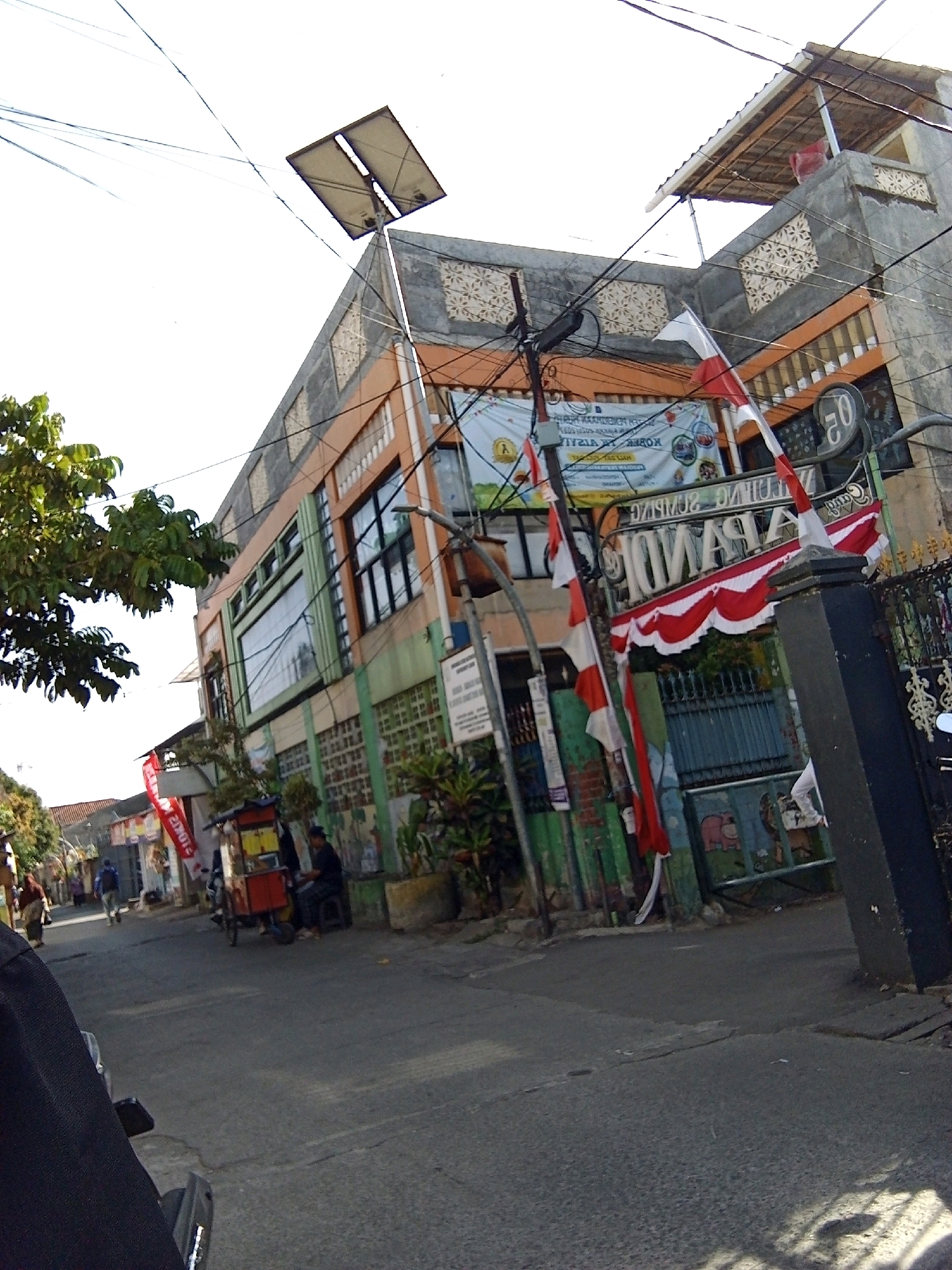 The image captures a street scene in Bandung City, Indonesia, likely in a commercial or residential area. The foreground features the left side of a black vehicle, suggesting the viewer is on a motorcycle or in a car. A paved road occupies the lower portion of the frame, with some shadows cast on it.
Dominating the midground and background is a multi-story building with a distinctive facade. The building's exterior is a mix of orange and green paint, with several windows visible, some reflecting the sky. Several Indonesian flags, red and white, are prominently displayed, attached to poles and draping down. One flag is tied to a lamppost with wires. A banner with text, though largely illegible, is also attached to the lamppost.
A utility pole with numerous wires runs across the scene, with a solar panel attached to the top of one of its extensions. The building also has decorative elements on its upper stories, including patterned square panels.
On the street in front of the building, a red and yellow food cart is visible, with a person sitting on a dark stool next to it. Further down the street, other individuals can be seen walking or standing. Trees and foliage are present on the left side of the image, adding a touch of greenery to the urban landscape. The overall impression is of a bustling street with a mix of everyday activities and nationalistic symbols.