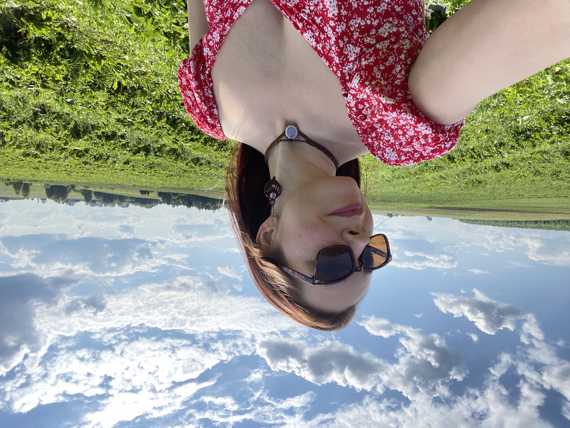 A woman with auburn hair, wearing sunglasses, a red floral print dress, a macrame choker with a light blue stone, and macrame earrings, stands in a field of green crops under a partly cloudy blue sky. The location is the village of Foteni, Russia.