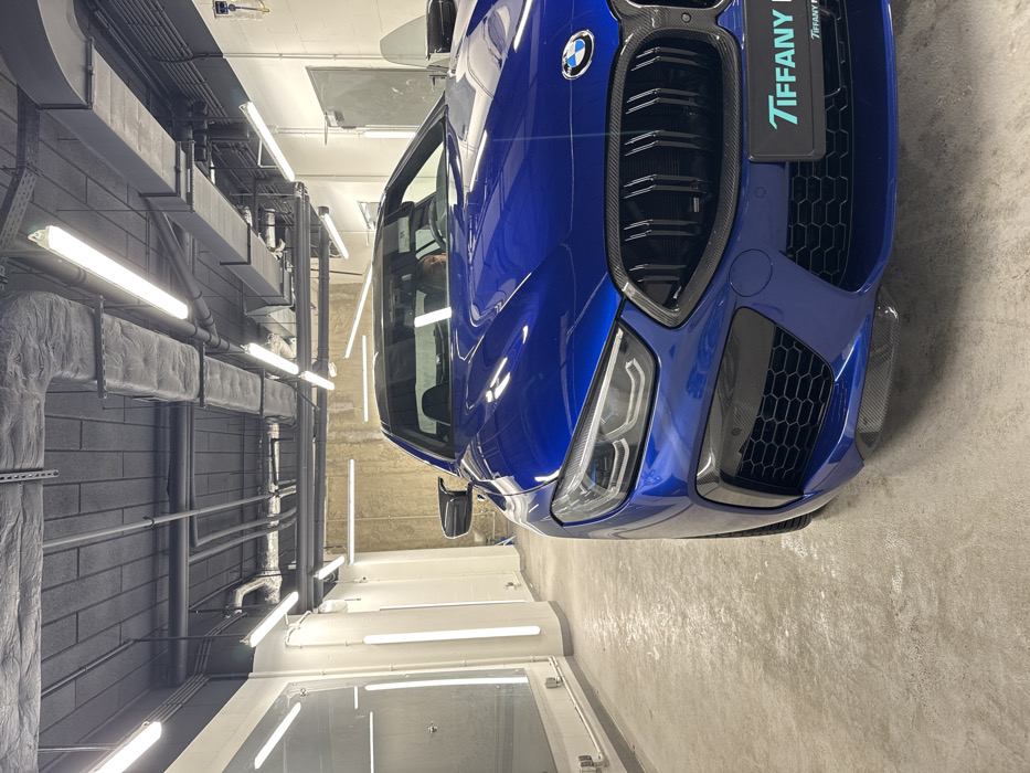 A dark blue BMW M8 Competition Coupe is centrally positioned indoors within an automotive detailing facility identified as Tiffany Premium Detailing, located at Al. Jerozolimskie 200, 02-486 Warsaw, Poland. The vehicle faces slightly left towards the viewer, featuring a prominent black kidney grille with vertical slats, LED headlights, and carbon fiber elements on the front bumper and lower splitter. A license plate holder on the front bumper displays "TIFFANY PR" and "Tiffany Premium Detailing". A person is partially visible in the driver's seat, showing only the upper torso and head with dark hair.
The environment consists of a light gray concrete floor and a dark gray industrial ceiling with exposed utility pipes, ducts, and numerous linear fluorescent light fixtures providing bright, even illumination. The left side of the scene features a white wall with vertical lighting elements and two clear, reflective panels. The background on the right displays a textured concrete wall and a closed gray metal door with a small rectangular window. The facility appears meticulously clean and well-lit, typical of a professional vehicle care establishment.