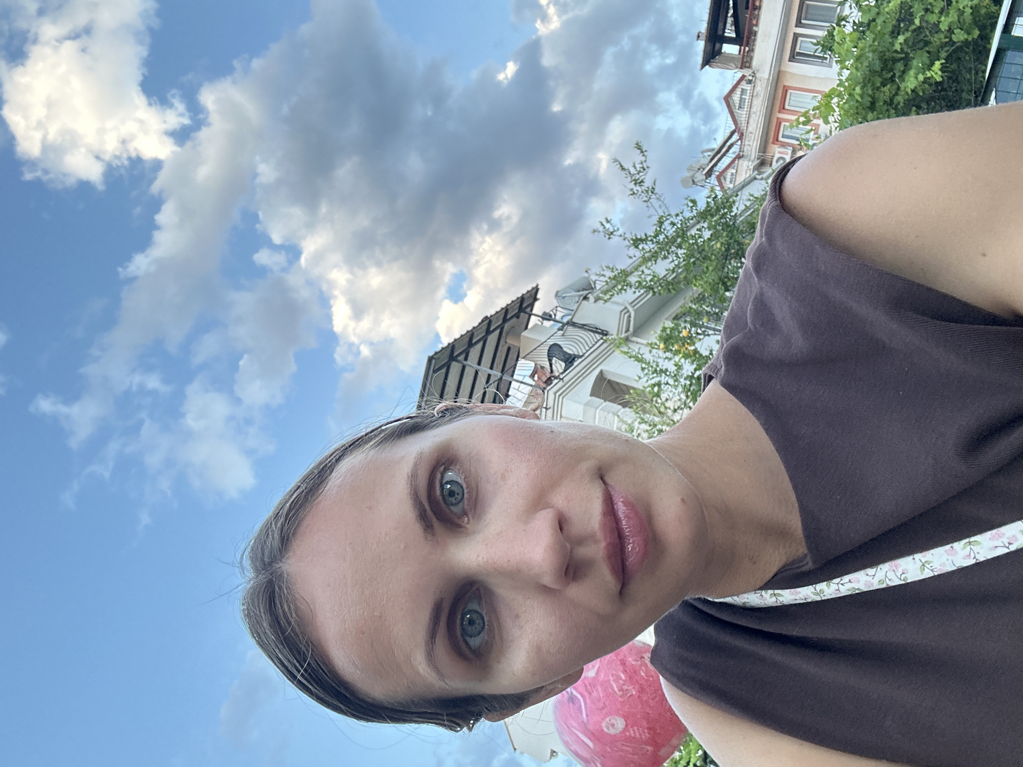A selfie shows a fair-skinned woman in a brown sleeveless top with a white floral-patterned strap, presumably taken outdoors in Fethiye, Turkey. The view is directed upwards, revealing a blue sky with scattered white and grey clouds. Behind the woman, a multi-story building with white walls and brown window frames is partially visible, along with some green trees. A pink object, likely a balloon or similar item, appears partially in the lower frame. The scene suggests an urban setting on a clear day.