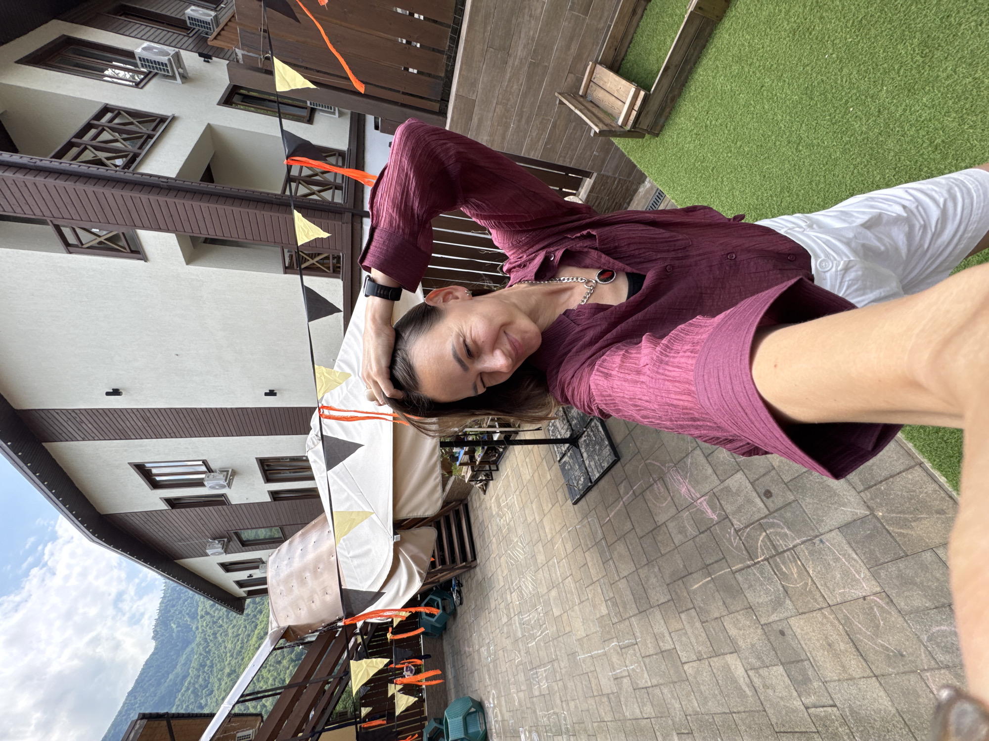A woman with dark hair is taking a selfie outdoors in a paved courtyard in Krasnaya Polyana, Russia. She is wearing a dark red crinkled blouse and white pants. She has her right arm raised and is touching her hair, with her eyes closed and a smile on her face. She is wearing a silver necklace with a red pendant. In the background, there is a multi-story building with balconies and air conditioning units, and mountains covered in green trees. Decorative flags are strung across the courtyard. There is also a sandbox with wooden borders on a patch of artificial grass. The paving stones of the courtyard are marked with chalk drawings.