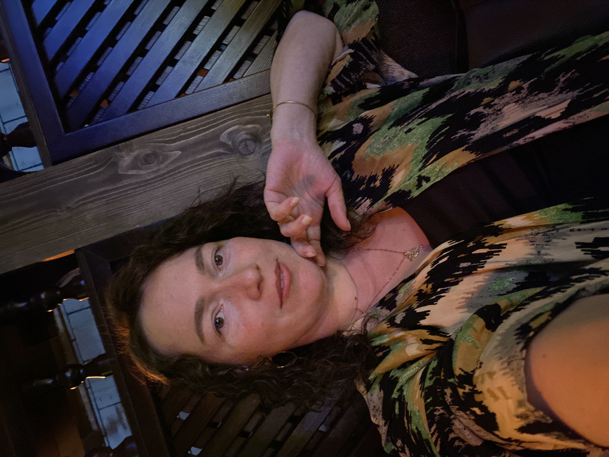 A medium shot depicts a woman reclining in an outdoor setting. Her face is centered, and she looks directly at the camera. She is fair-skinned with brown eyes and dark curly hair. Her left hand is raised near her face. She wears a patterned dress in shades of green, yellow, and black, along with a gold necklace and bracelet. The scene is set on a wooden structure with a slatted design. The location is not discernible from the image.