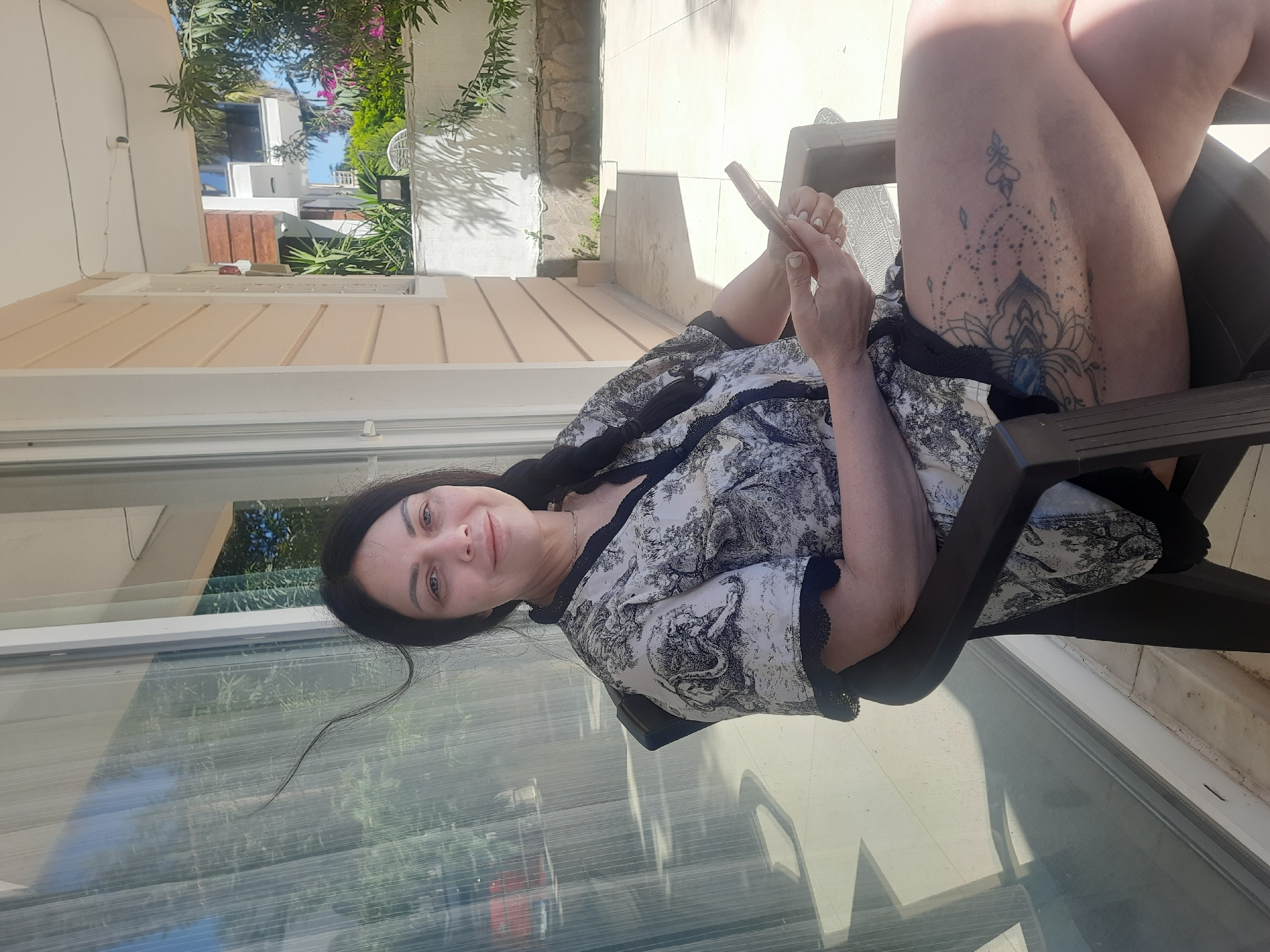A woman is sitting in a dark chair on a patio, using a phone. She has long dark hair. She is wearing a patterned black and white top. The woman has a tattoo on her upper right thigh. The patio has a glass wall. Outside, there is vegetation and a building. This scene is likely in Kemer, Turkey.