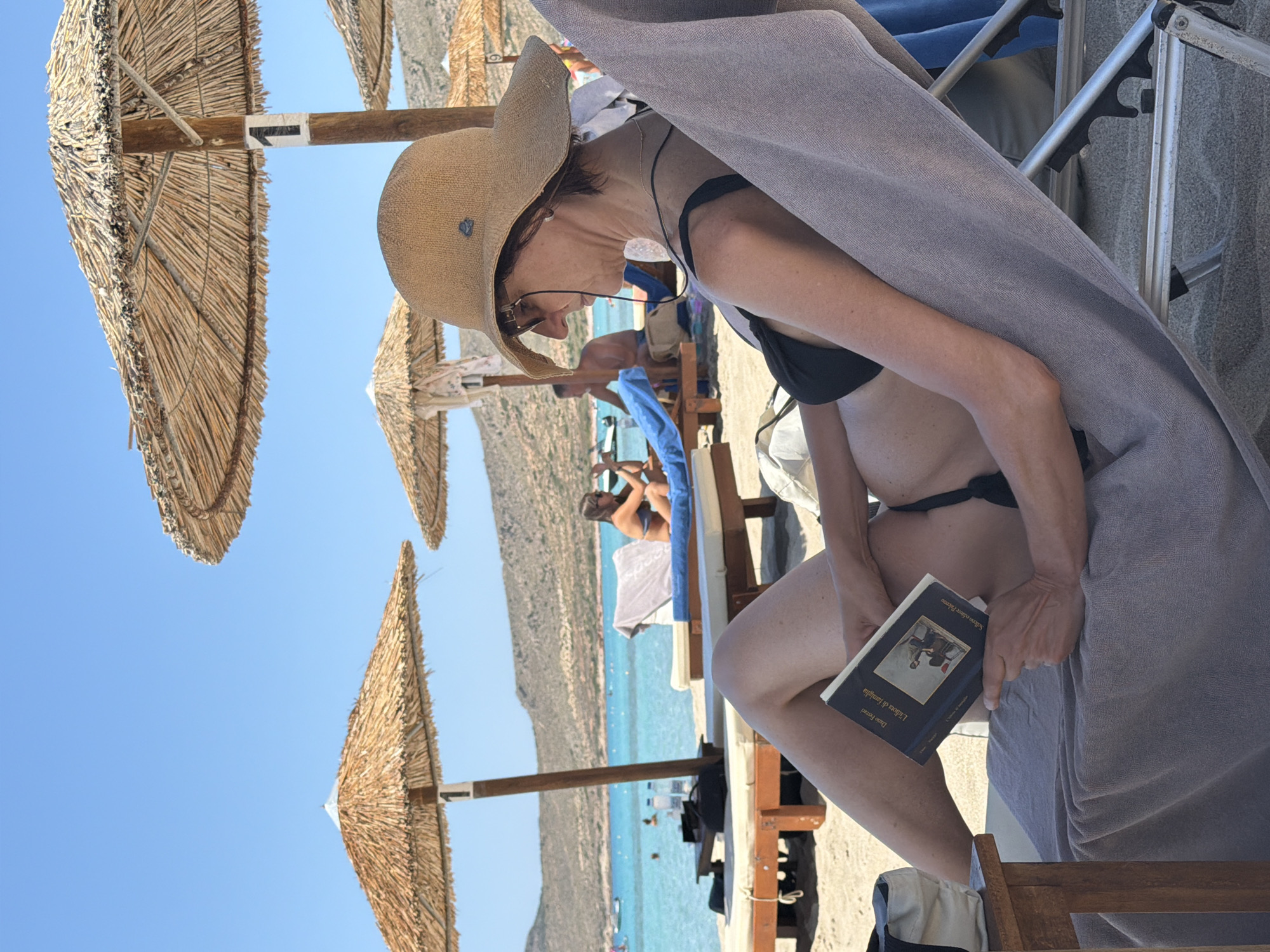 The image captures a woman wearing a wide-brimmed straw hat and sunglasses, dressed in a black bikini top, relaxing on a beach chair on a sunny day. She is holding and reading a book with a dark cover and a picture on it. The beach chair is covered with a gray towel. In the background, there are several thatched umbrellas providing shade, with the sea and a coastline with hills visible beyond. Other beachgoers can be seen in the distance, some on lounge chairs and others near the water. The scene is set on a sandy beach with clear turquoise water and a bright blue sky. The location is identified as a beach in Greece.