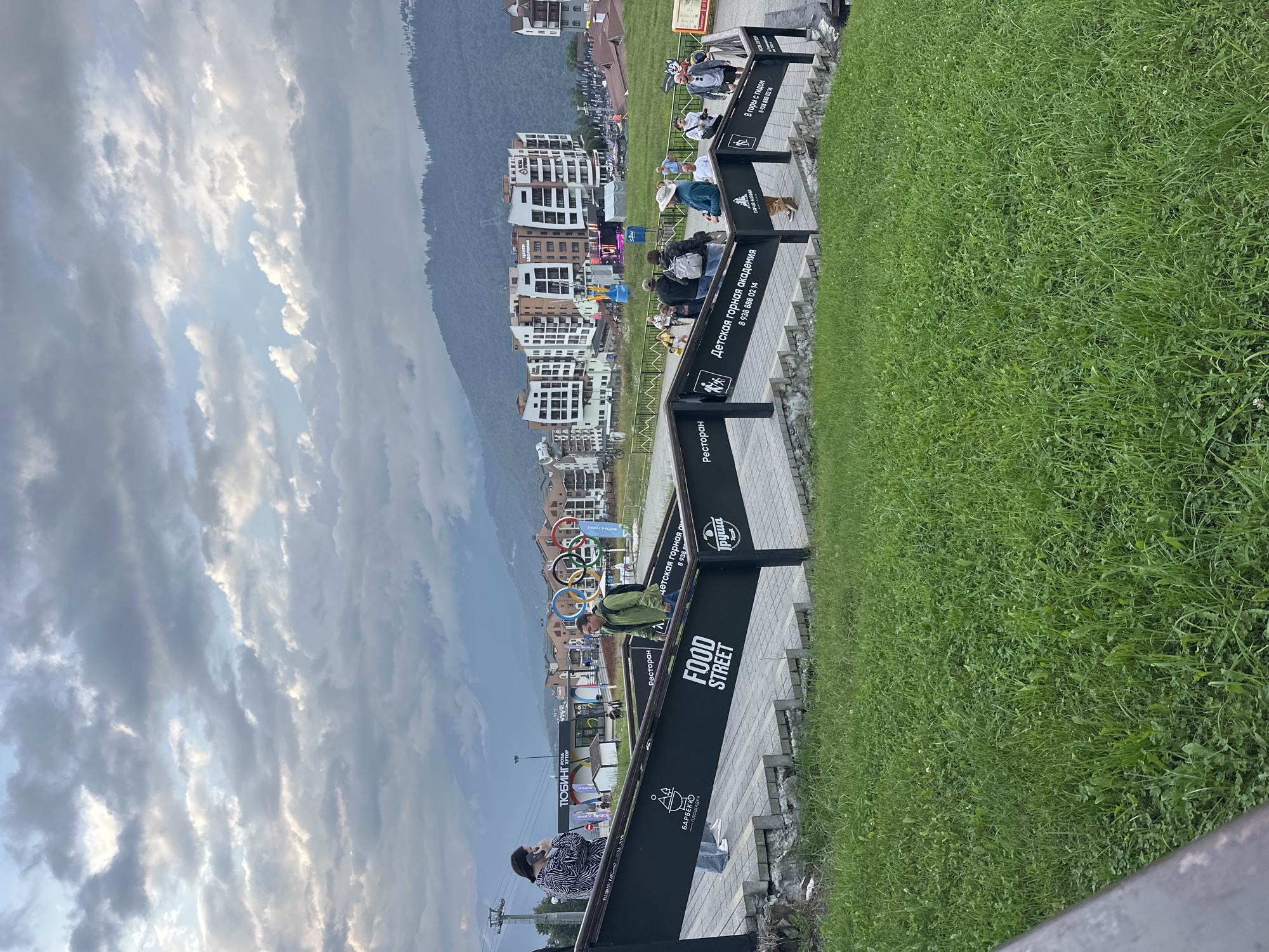 The image shows a view of the Esto-Sadok district in Sochi, Russia, on a cloudy day. In the foreground, a grassy slope leads up to a set of stairs with black railings. Signs on the railings indicate "FOOD STREET" and "BARBECUE," as well as "Restaurant" and "Children's Mountain Academy." Several people are walking up and down the stairs. In the middle ground, there are buildings with modern architecture, including what appears to be a resort or residential complex. Olympic rings are visible in the background, near the buildings. Mountains covered with trees form the backdrop of the scene. The sky is filled with dramatic, overcast clouds.