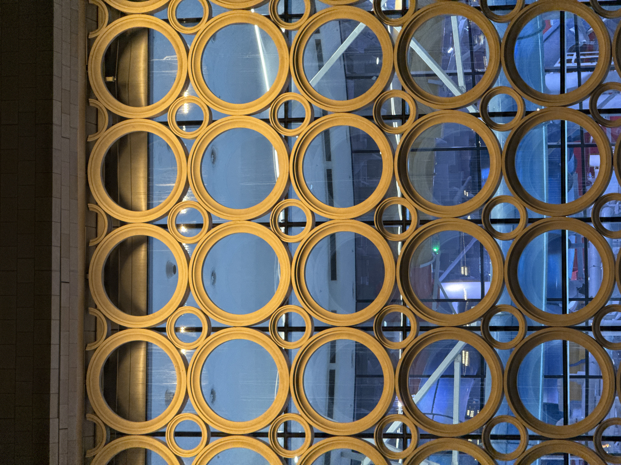 The image shows a geometric window design composed of interconnected gold-colored circles of varying sizes. Through the circles, a blue-toned interior is visible, showing architectural elements such as structural beams, lights, and windows. The left side of the image includes a portion of a brick or tile wall. This design is located at the Zaryadye Park in Moscow, Russia.