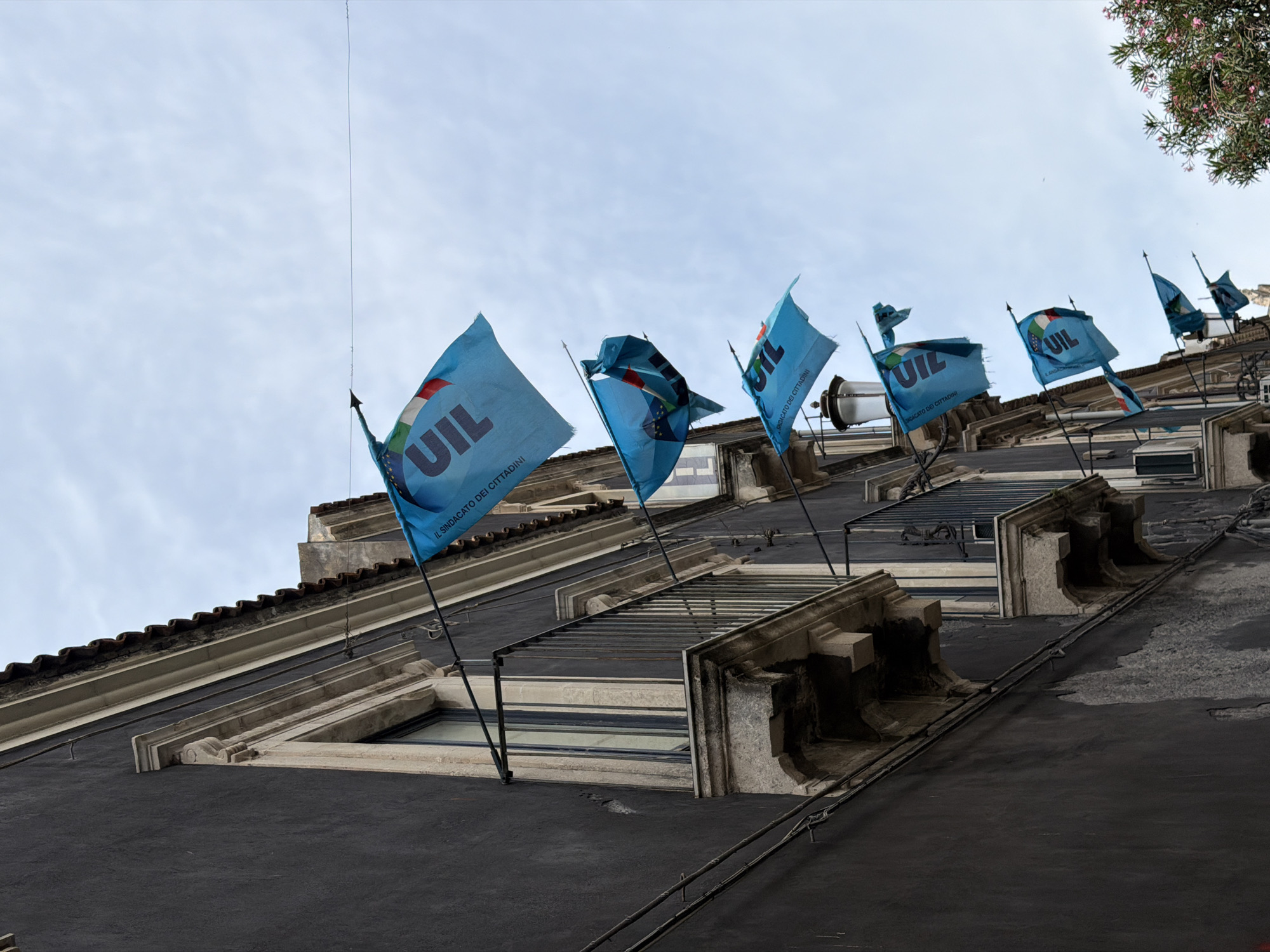 The image captures the facade of a building in Catania, Italy, adorned with a series of light blue flags bearing the "UIL" acronym. These flags, waving in the wind, are attached to poles mounted on the building's exterior. The building itself has a dark, textured facade, with protruding window ledges and decorative architectural details. The sky is overcast. Some trees are visible in the upper right corner of the image.