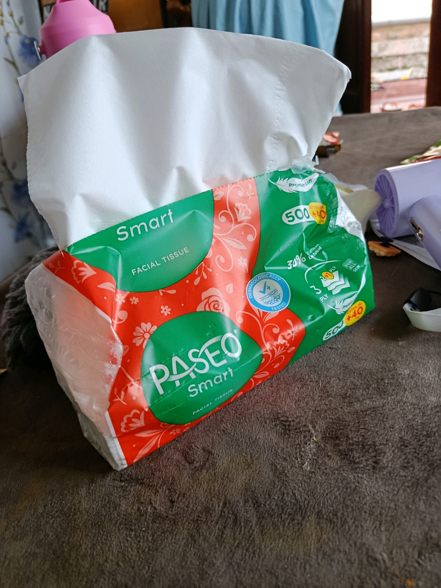The image shows a pack of Paseo Smart Facial Tissues on a textured, dark brown surface, possibly a table or sofa. The tissue pack has a green and orange design with floral motifs. White tissues are visible peeking out from the top of the pack. In the background, a pink object, possibly a bottle, is partially visible on the left, and a light blue fabric is visible on the upper right. A blurred background suggests an indoor setting with some light coming from the right. No people are present in the image. The location of Karang Rejo, Indonesia is not depicted or suggested in the image itself.