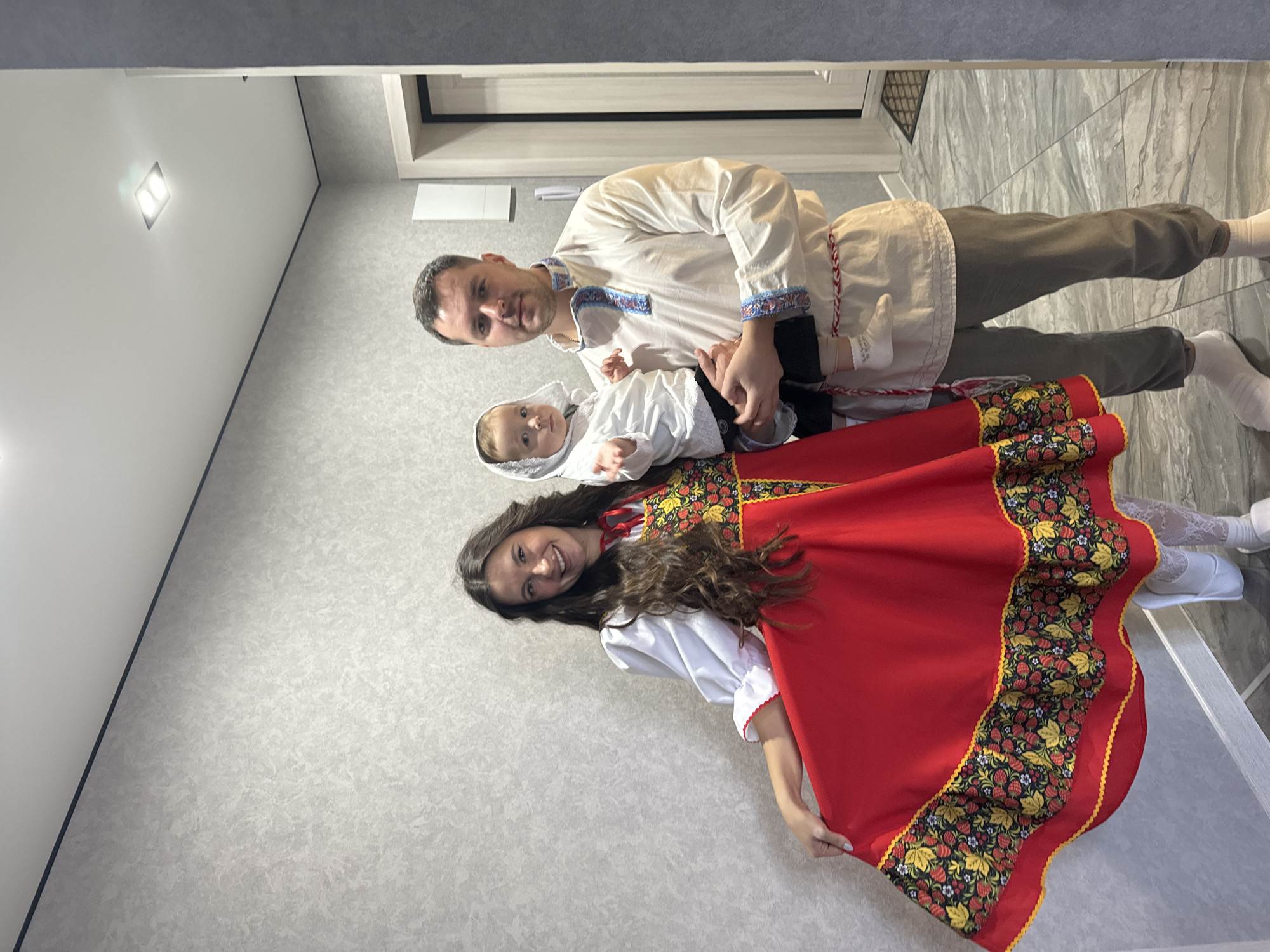 A family of three are posing indoors. The father, wearing a traditional white shirt with embroidered blue and red details, is holding a baby, who is also dressed in white. To the left of the father and baby stands a woman wearing a red dress with a patterned trim, the dress is paired with a white blouse underneath. She is smiling and holding up the edge of her dress. They are standing against a light grey patterned wall, in what appears to be an apartment hallway or entrance area, somewhere in the city of Unknown, Unknown. A doorway can be seen in the background with a white door frame.
