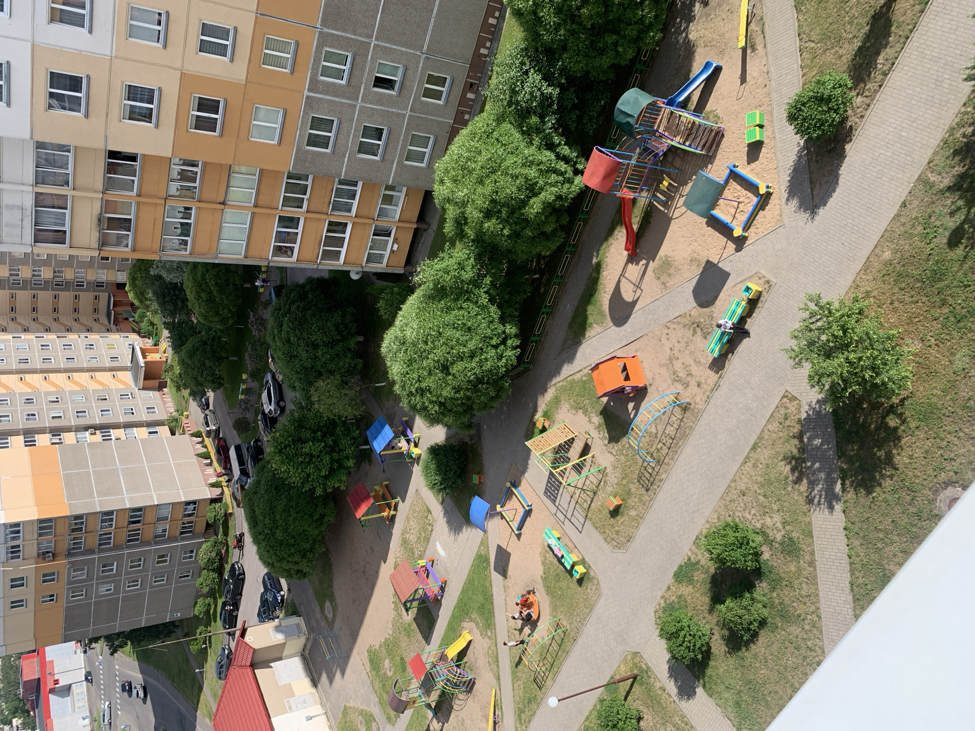 This high-angle, aerial view depicts a residential area within the city of Hrodna, Belarus, under clear, sunny conditions with distinct shadows. The scene encompasses apartment buildings, a street, parking areas, and a central children's playground.
In the background, a multi-lane asphalt road is visible, with several vehicles, including sedans and a motorcycle, either driving or parked. Further back on the left, a large red-roofed commercial building or warehouse can be seen. In the mid-ground to the left, a prominent multi-story apartment building features a grey lower section and orange/yellow upper facades with numerous windows and balconies. Adjacent to it, a smaller, single-story building with a red tiled roof and beige walls stands out. To the right, another multi-story apartment building, primarily beige/orange and white with grey base sections, also features multiple windows.
The central and foreground areas are dominated by a children's playground constructed on a mix of sand and paved pathways. The playground contains diverse colorful equipment, including: multiple climbing structures with slides (one yellow, one blue with a wooden ramp), two small gazebo-like playhouses (one red-roofed, one blue-roofed), a blue-canopied sandbox, a circular spinning carousel (red and yellow), various climbing frames (metal, triangular wooden, arched blue), benches (green and yellow), and a yellow seesaw.
Several individuals are present in the playground: three adults and one child are gathered around the red and yellow carousel, one person is seated on a green and yellow bench near the blue sandbox, and other figures are vaguely discernible near play structures. The entire area is interspersed with green spaces, including mature trees with full foliage, various bushes, and grassy areas, some of which appear dry. Cars are parked along the street and in informal parking areas near the apartment buildings.