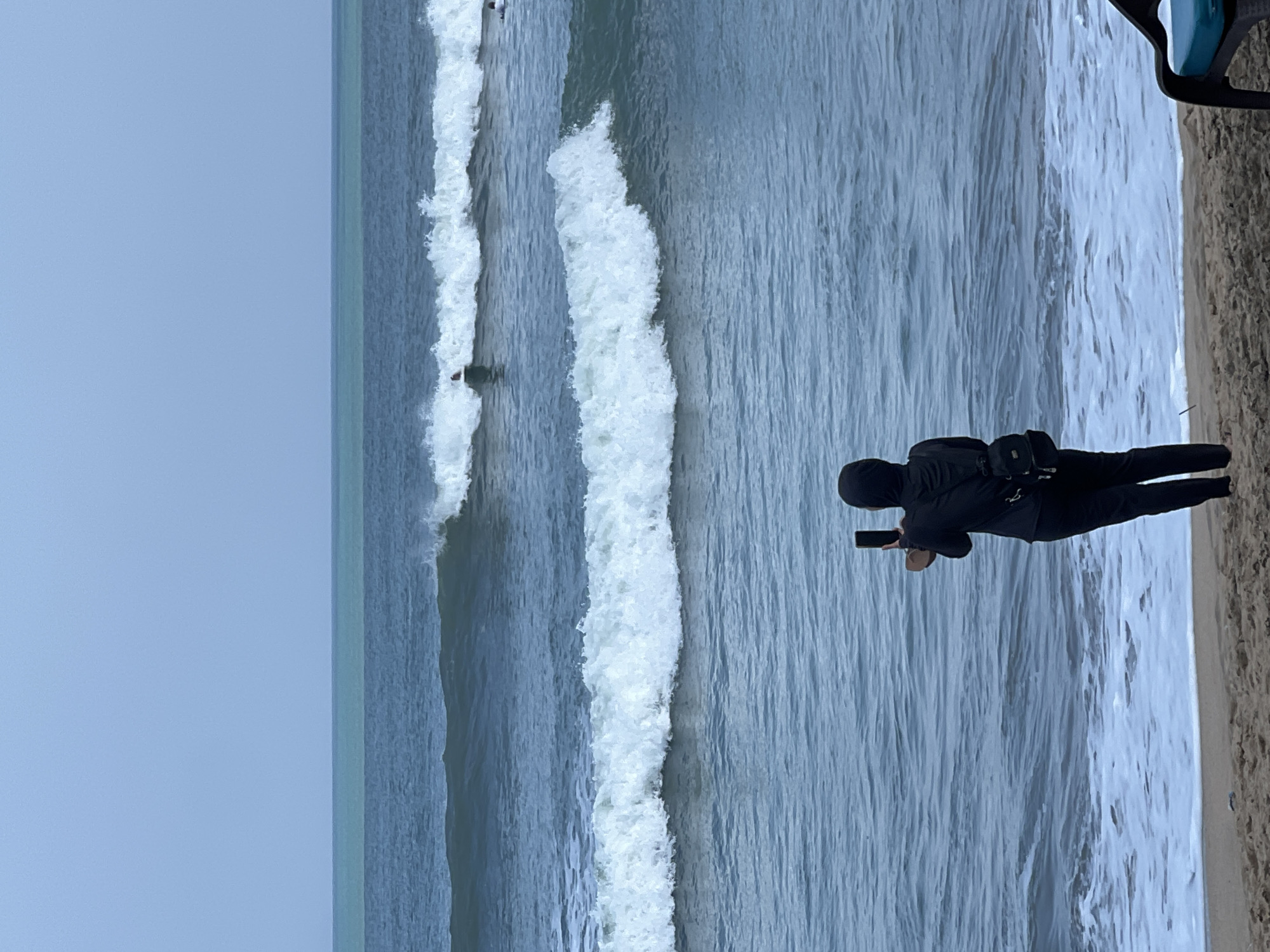 A person wearing a black hijab and black athletic clothing stands on a sandy beach in Kuta, Indonesia, facing the ocean. They are holding a mobile phone in their right hand, seemingly taking a picture or video of the waves. A small black backpack is visible on their back. The ocean is choppy with several waves breaking, some of which have white crests. In the distance, two surfers are visible on their surfboards, riding or preparing to ride waves. The sky is overcast with a pale blue-grey hue. The scene captures a moment of observation and engagement with the coastal environment.