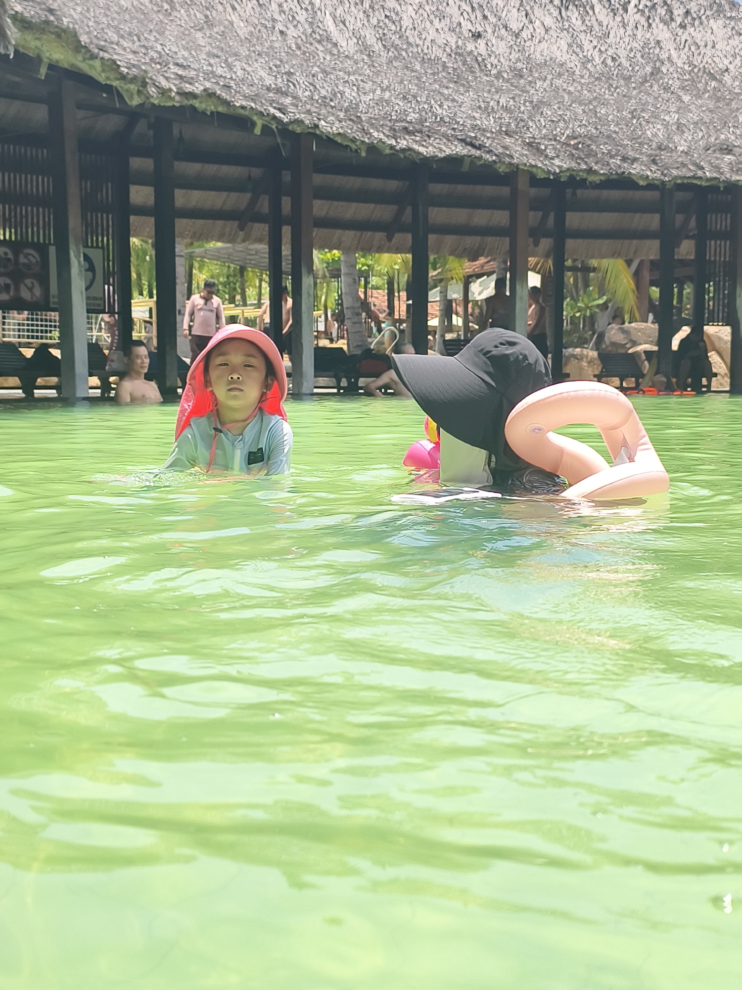 The image depicts two individuals in a body of water in Tay Nha Trang Ward, Vietnam. The foreground shows a child wearing a hooded pink swimsuit. Behind them, partially submerged, is an adult wearing a wide-brimmed black hat and a peach-colored inflatable ring around their neck. The water appears to be light green. Behind the subjects, a covered structure with wooden pillars and a thatched roof is visible. In the background, figures and some seating are seen under the shelter.