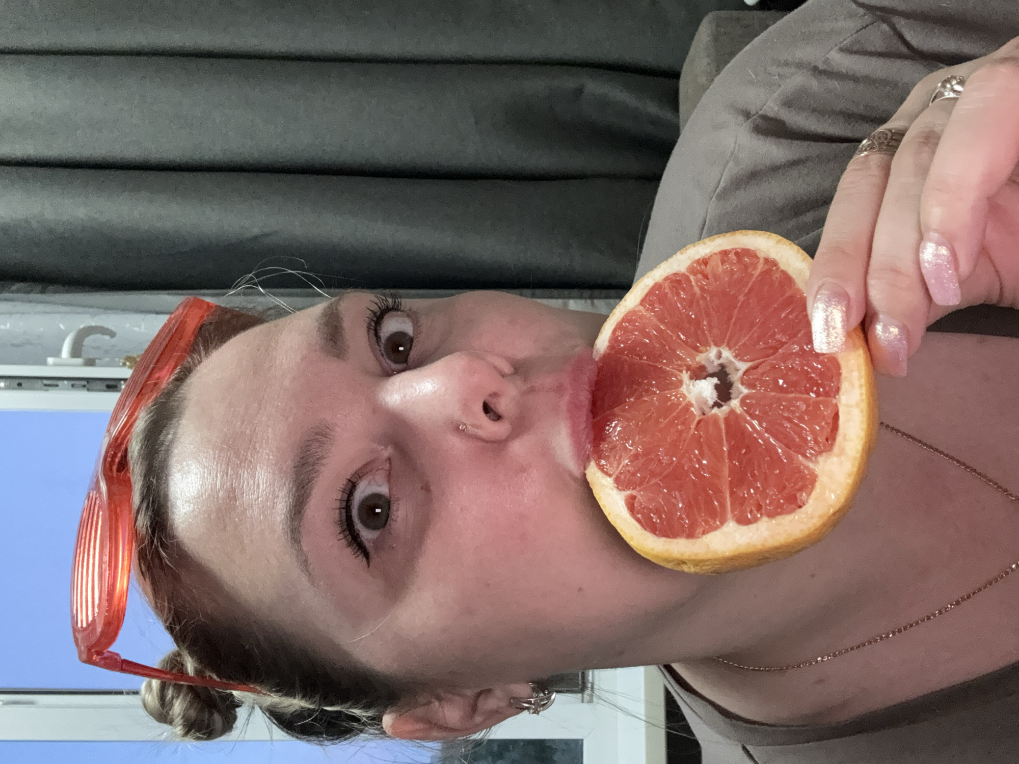 A woman is holding a grapefruit up to her mouth. She has her hair in a bun, wears glasses, and has fingernails with pink glitter polish. The background shows curtains, and a window with blue sky visible.