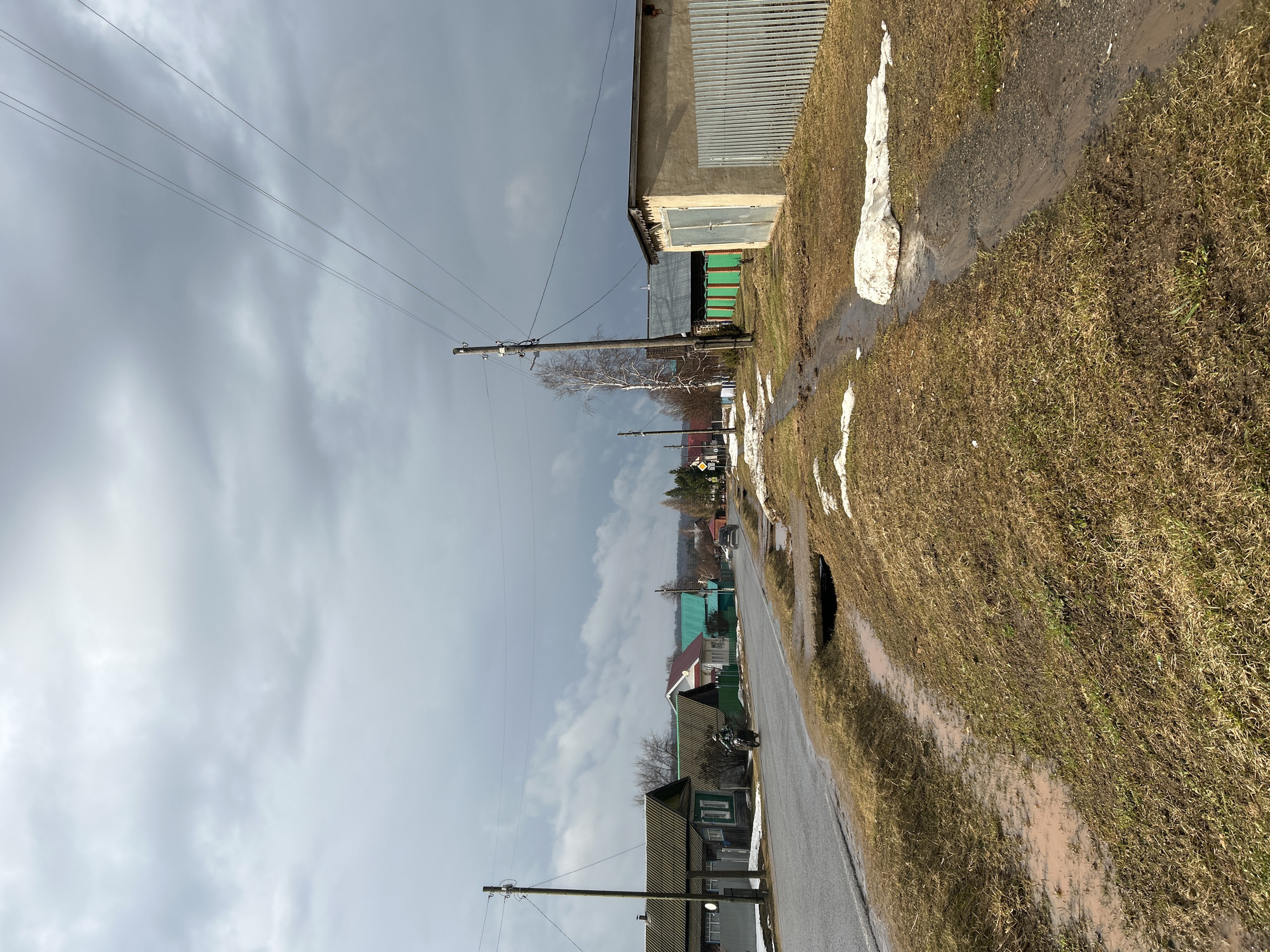 The image depicts a street scene in Уральское, Russia. The viewpoint is from the side of a wet asphalt road, alongside which lies a strip of grass and mud, with patches of melting snow. A ditch parallels the road, carrying runoff. Beyond the road are houses, some with green roofs. Utility poles run along the street, with power lines spanning overhead against a gray, overcast sky. In the distance, other buildings and vegetation are visible. A parked motorcycle is visible along the road. The architecture of the buildings is typical of rural Russian settlements.