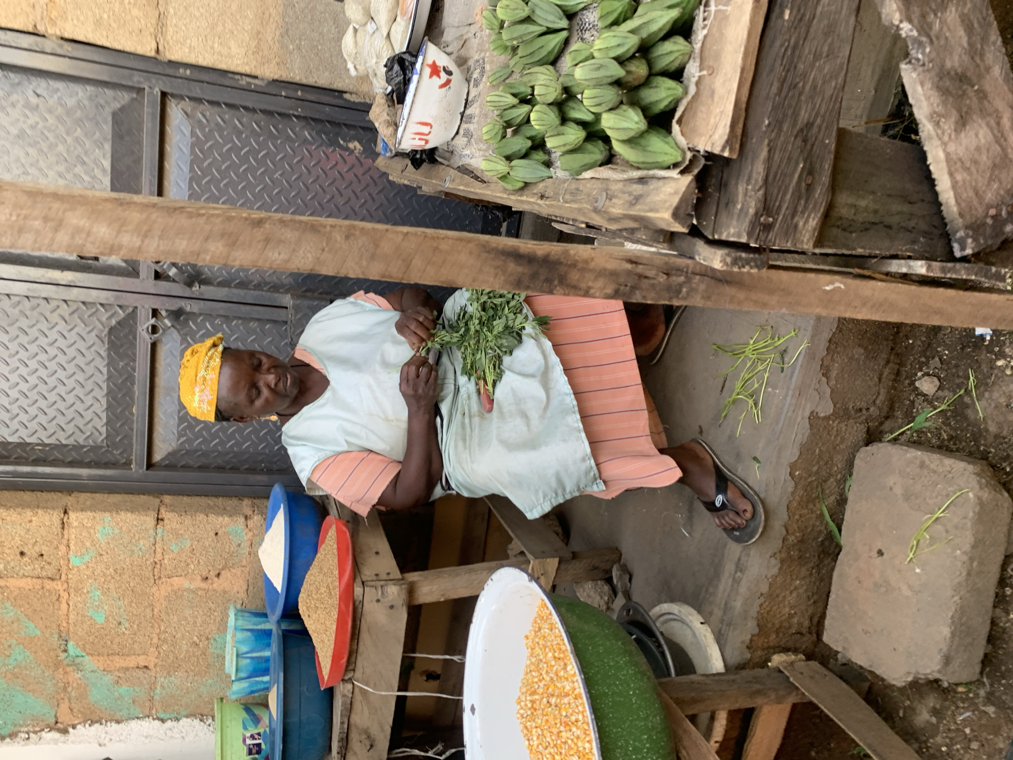 The image shows an elderly woman in Jos, Nigeria, sitting outdoors at a market stall. She wears a yellow headscarf, a light-colored top, and a striped skirt. She is holding a bundle of green leafy vegetables in her hands. In front of her are several bowls and containers filled with different grains and seeds, possibly for sale. Beside her is a pile of green okra in a wooden crate. The background includes a metal door and a brick wall. The ground around her is uneven and appears to be a dirt and concrete mix.