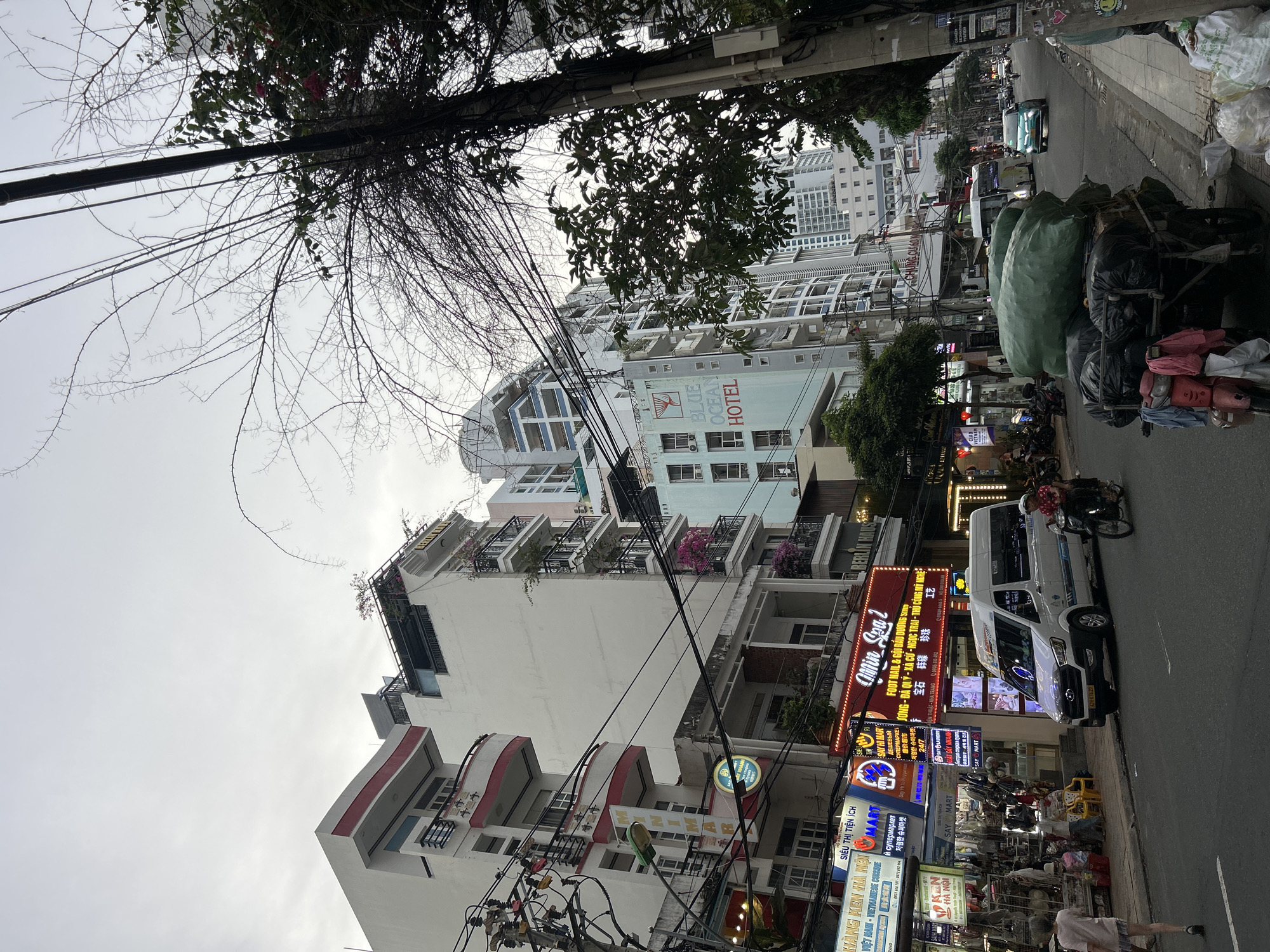 This image depicts a vibrant street scene in Nha Trang Ward, Vietnam, captured from a low angle looking upwards towards the buildings under an overcast sky. The street is lined with multi-story buildings, some appearing residential with balconies adorned with plants, and others housing commercial establishments. A prominent "BLUE OCEAN HOTEL" sign is visible on a light blue building with distinctive arched window designs.
The scene is bustling with urban activity. A white van is parked or driving along the street, and a person is riding a motorcycle. Another individual is seen walking on the sidewalk. A key detail is a heavily loaded pushcart or motorcycle trailer, piled high with large green and black bundles, suggesting goods delivery or waste collection. Numerous electrical and telephone wires crisscross overhead, characteristic of Vietnamese cityscapes, with various trees, some bare-branched and some leafy, intertwined with them.
The street level is illuminated by numerous brightly lit, colorful shop signs advertising a variety of businesses. Visible text includes:
* "BLUE OCEAN HOTEL"
* "SIÊU THỊ TIỆN ÍCH" (Convenience Supermarket)
* "HÀNG KEN HÀ NỘI" (Hanoi Ken Goods/Shop) with "VIỆT NAM - VIETNAMESE CORSIDE"
* "SAY MART"
* "Min Spa 2" offering "FOOT NAIL & GỘI ĐẦU DƯƠNG SIMP" (Foot Nail & Hair Wash) along with mentions of gems, clams, pearls, and crafts.
* Translations for "Cheap Supermarket" in Russian ("DEШЕВЫЙ СУПЕРМАРКЕТ"), Korean ("저렴한 슈퍼마켓"), and Chinese ("廉价超市").
* "SAY LAUNDRY GIẶT SẤY NHANH" (Quick Wash & Dry).
The presence of diverse languages on the signs caters to international visitors, indicating a tourist-friendly area. The scene portrays a lively commercial district during what appears to be an overcast daytime or early evening, given the bright lights on some signs.