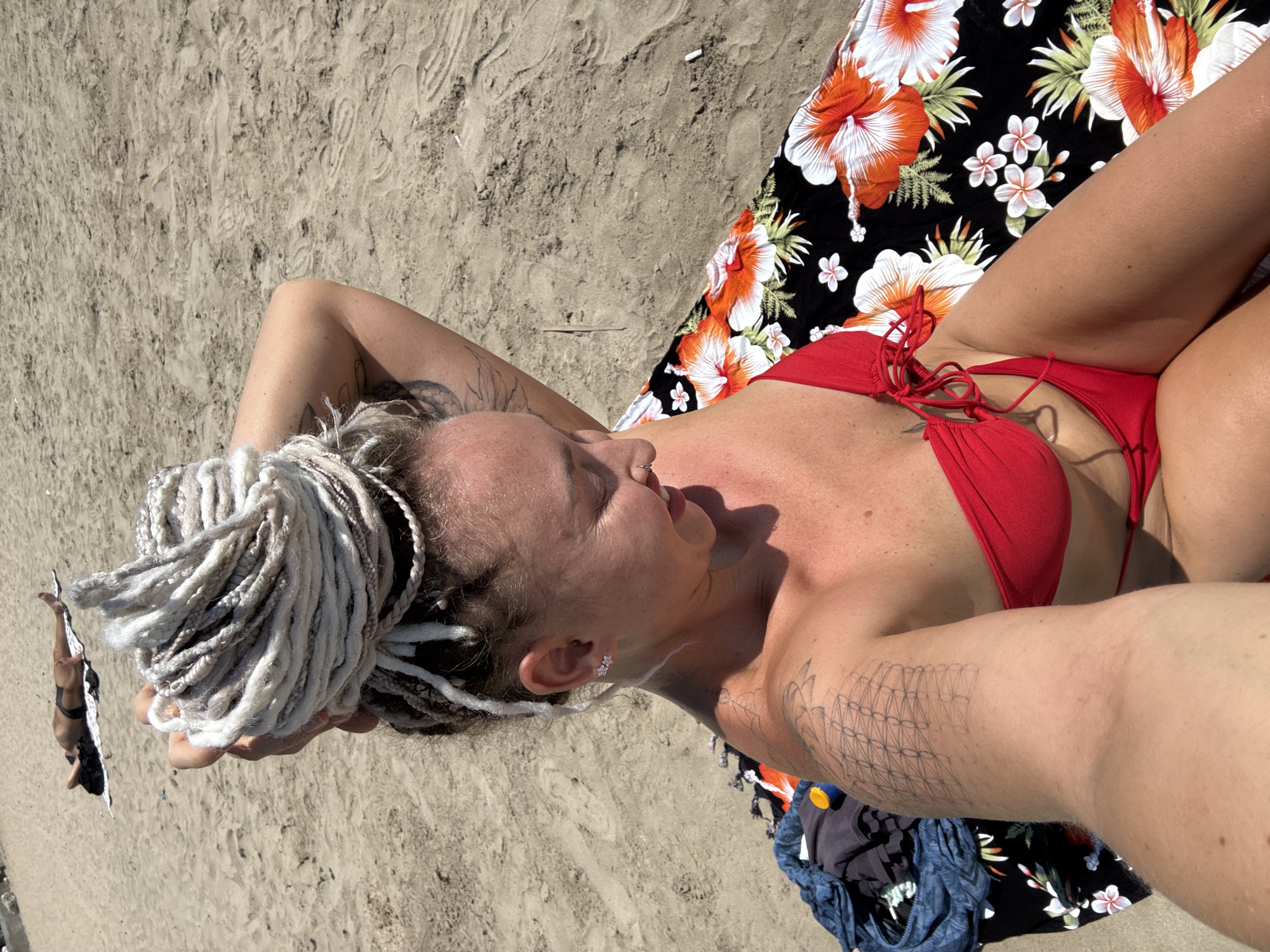 A woman with blonde dreadlocks styled in a bun is sitting on a beach towel with a floral pattern. She is wearing a red bikini and has tattoos on her arm and shoulder. She is looking down and smiling. In the background, another person is lying on the sand. The scene is a sandy beach. The location is the city of Bac Nha Trang Ward, Vietnam.