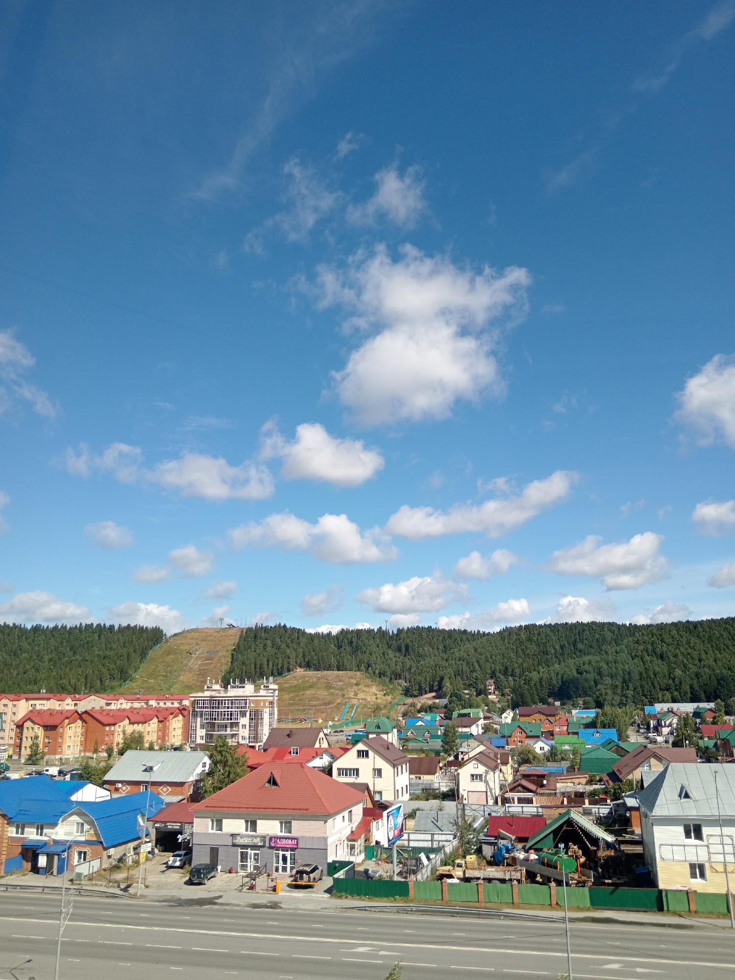 This is a high-angle, full shot of a residential area in Khanty-Mansiysk, Russia, under a bright blue sky dotted with white clouds. In the foreground, a paved road with white lane markings stretches horizontally. A light pole stands on the left side of the road. Behind the road, a collection of houses and buildings are visible, many with colorful roofs in shades of red, blue, and green. Some of these buildings appear to be multi-story apartment complexes with light-colored facades. Other structures are smaller, single-family homes. A few vehicles are parked in driveways or on the street. In the middle ground, a large, modern building with many windows stands out. Further in the background, a dense, green forest covers a sloping hill. On the side of the hill, a ski slope with ski lifts is discernible. The scene depicts a suburban or semi-rural environment with a mix of modern and traditional architecture.