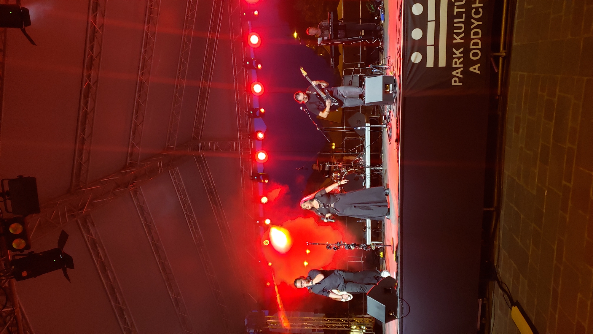 A band performs on an outdoor stage under a tent structure, illuminated by red stage lights. In the foreground, a male vocalist in a black shirt and pants stands to the left, holding a microphone in his right hand. To his right, a female vocalist with long pink hair, wearing a black dress with studded detailing, sings into a microphone. Behind them, a guitarist in a black shirt and pants plays an electric guitar, and further back, a drummer is partially visible behind a drum kit. To the far right, a keyboardist in a black shirt and pants plays a keyboard. The stage is set up on a paved area. A banner at the front of the stage reads "PARK KULTÚRU A ODDYCH". This scene is at the Culture and Relaxation Park (Park kultúry a oddychu) in Prešov, Slovakia.