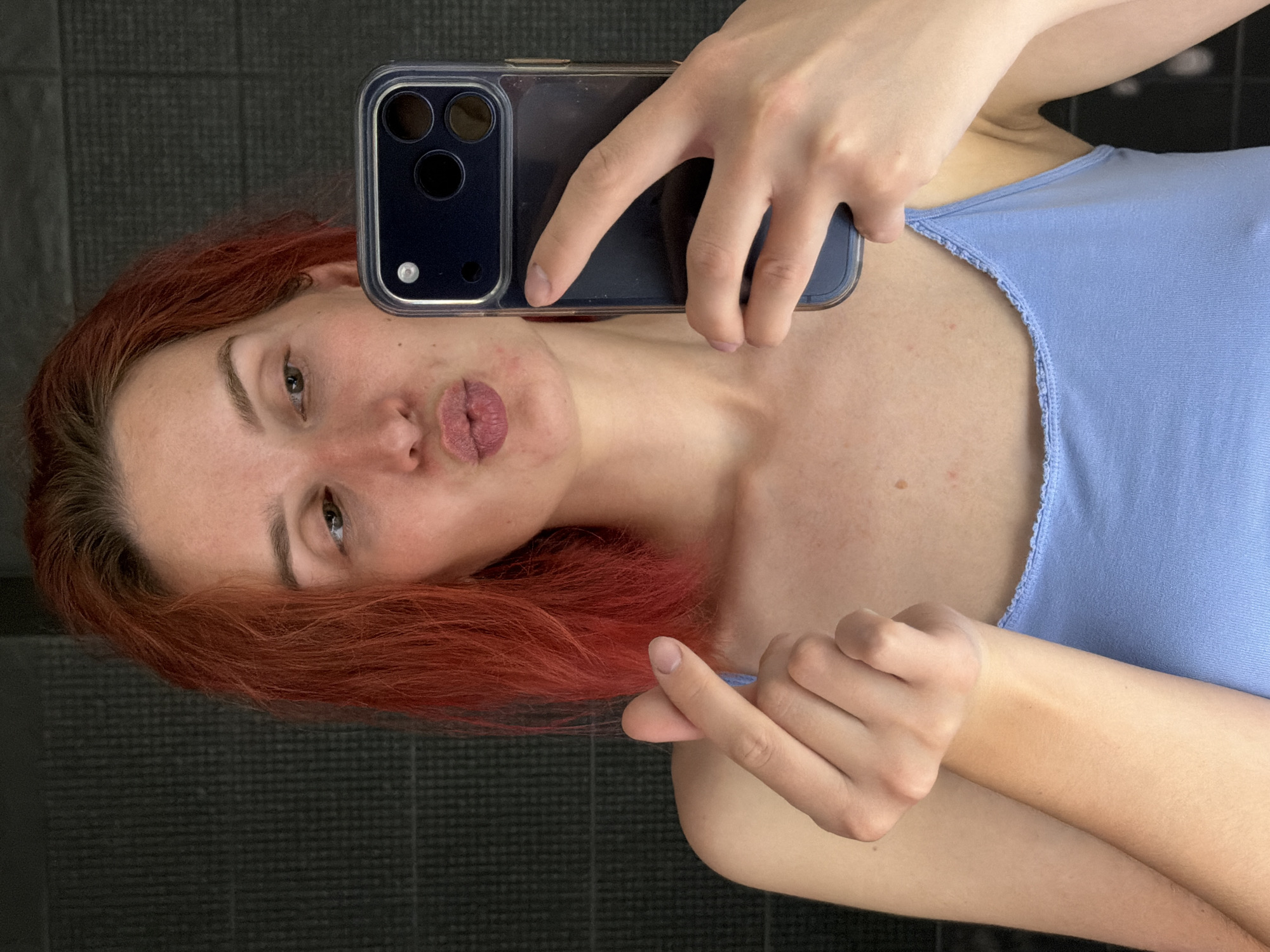 A selfie of a light-skinned person with red hair, wearing a blue tank top. The person is holding a smartphone with a black case. The background is a dark, possibly tiled wall. The location is unknown and cannot be determined from the image.