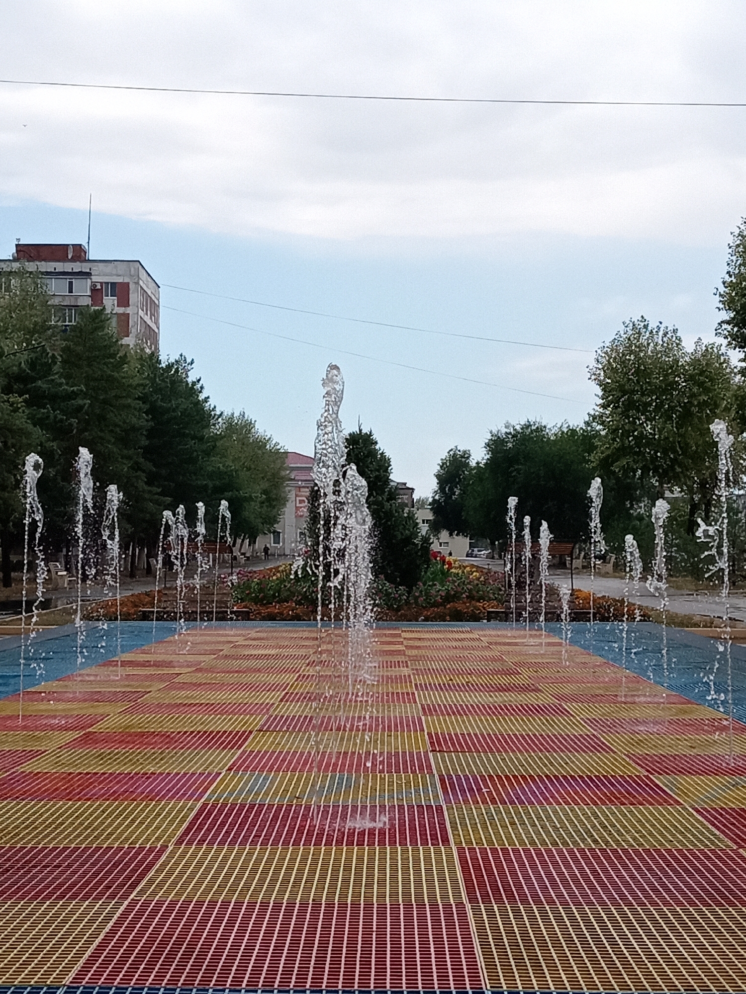 The image depicts a fountain in a public space in Rudny, Kazakhstan. The fountain is located on a checkered surface with red, yellow, and blue colors. Water jets are spraying upwards from the surface, creating a dynamic visual effect. In the background, there are trees, buildings, and a cloudy sky. The overall scene suggests a recreational area within the city.