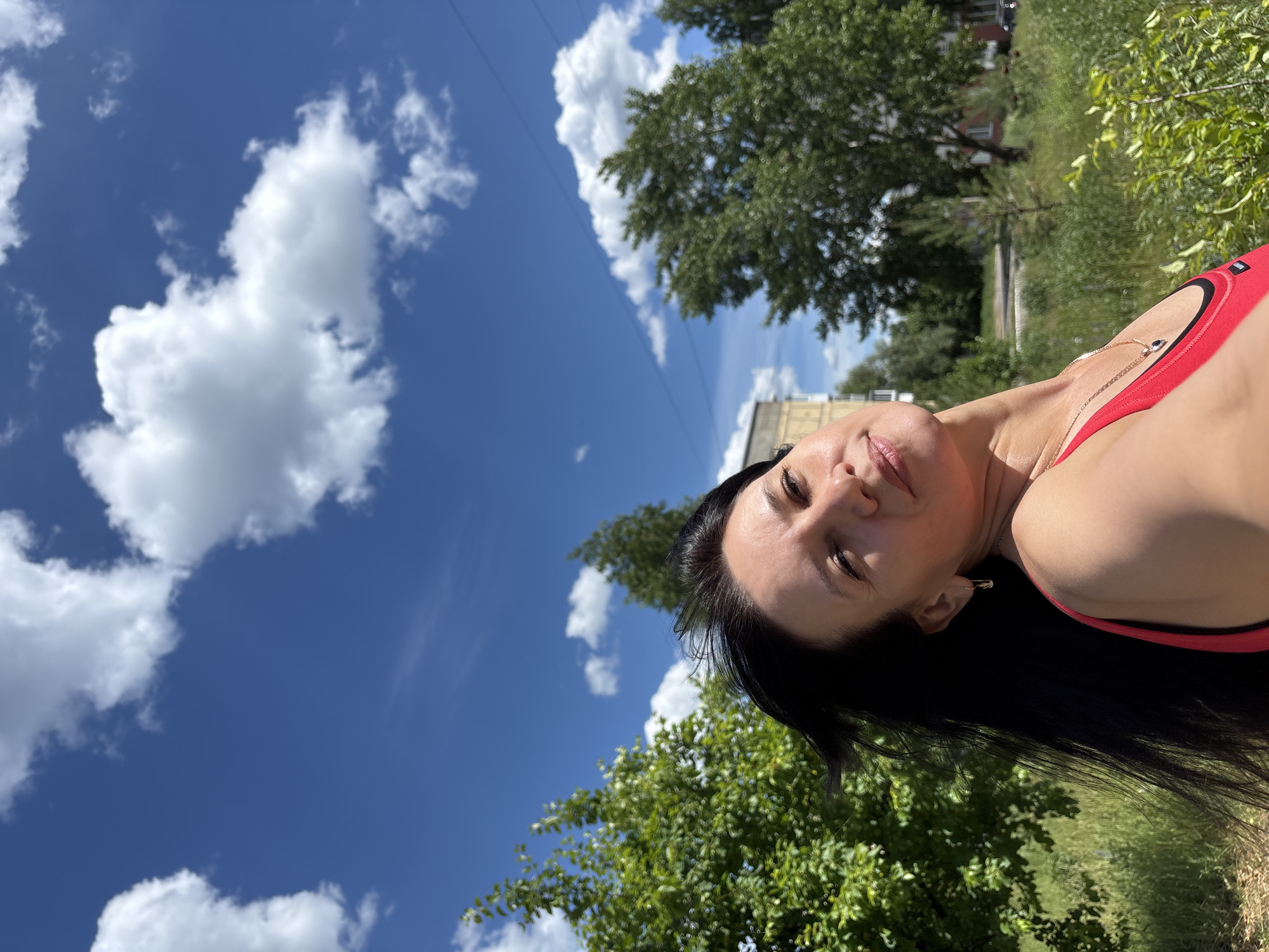 A young woman with long, dark hair is captured in a selfie outdoors. She is wearing a red top and a necklace. The background shows a blue sky with white clouds, green trees, and a building. The scene appears to be a residential area in the city of Chaikovsky, Russia, possibly a park or a street with vegetation. The lighting suggests it is daytime, with bright sunlight.