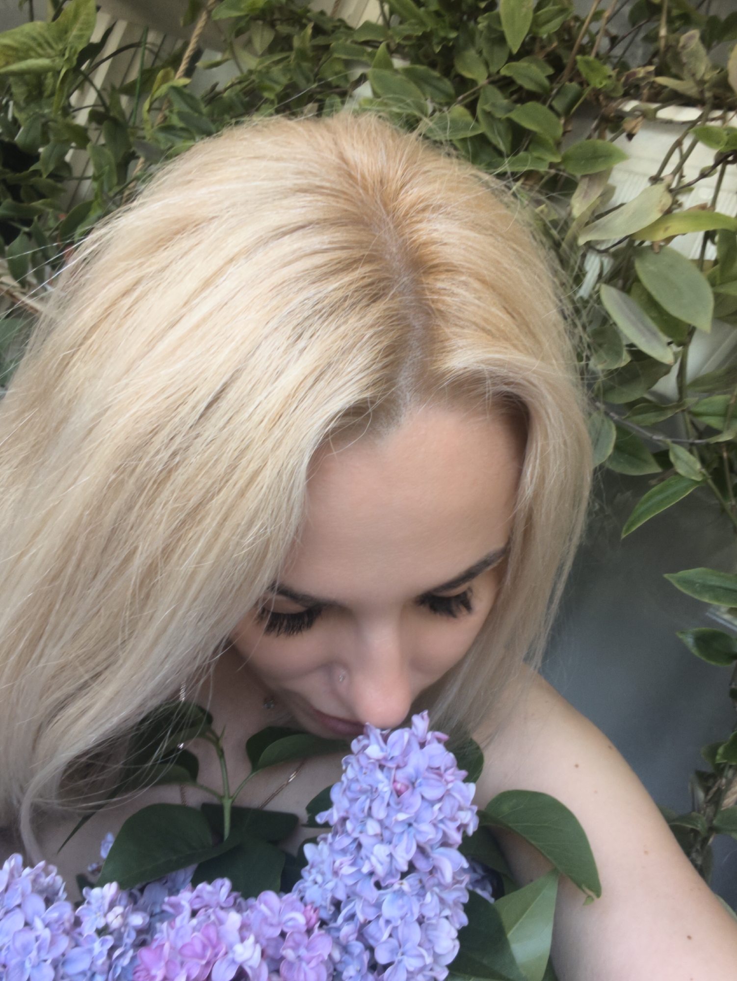 A light-skinned woman with long, blonde hair is smelling a bouquet of light purple lilacs. She has dark eyelashes and a piercing in her nose. She is positioned in front of foliage of green leafy plants. The location in the city of Sevastopol, Ukraine is not discernible from the image.