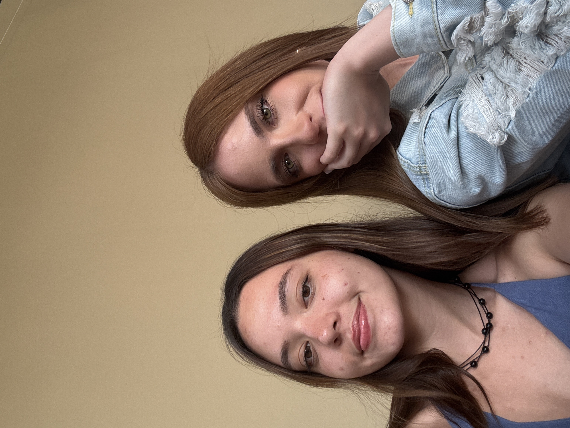 Two young women are captured in a selfie-style shot. The woman at the top has light brown hair, green eyes, and is wearing a distressed denim jacket. She has her hand near her face in a thoughtful pose. The woman at the bottom has darker brown hair, brown eyes, and is wearing a blue top and a black beaded necklace. The background is a plain beige wall. There is no information to suggest the image was taken in San Bernardino Tlaxcalancingo, Mexico.