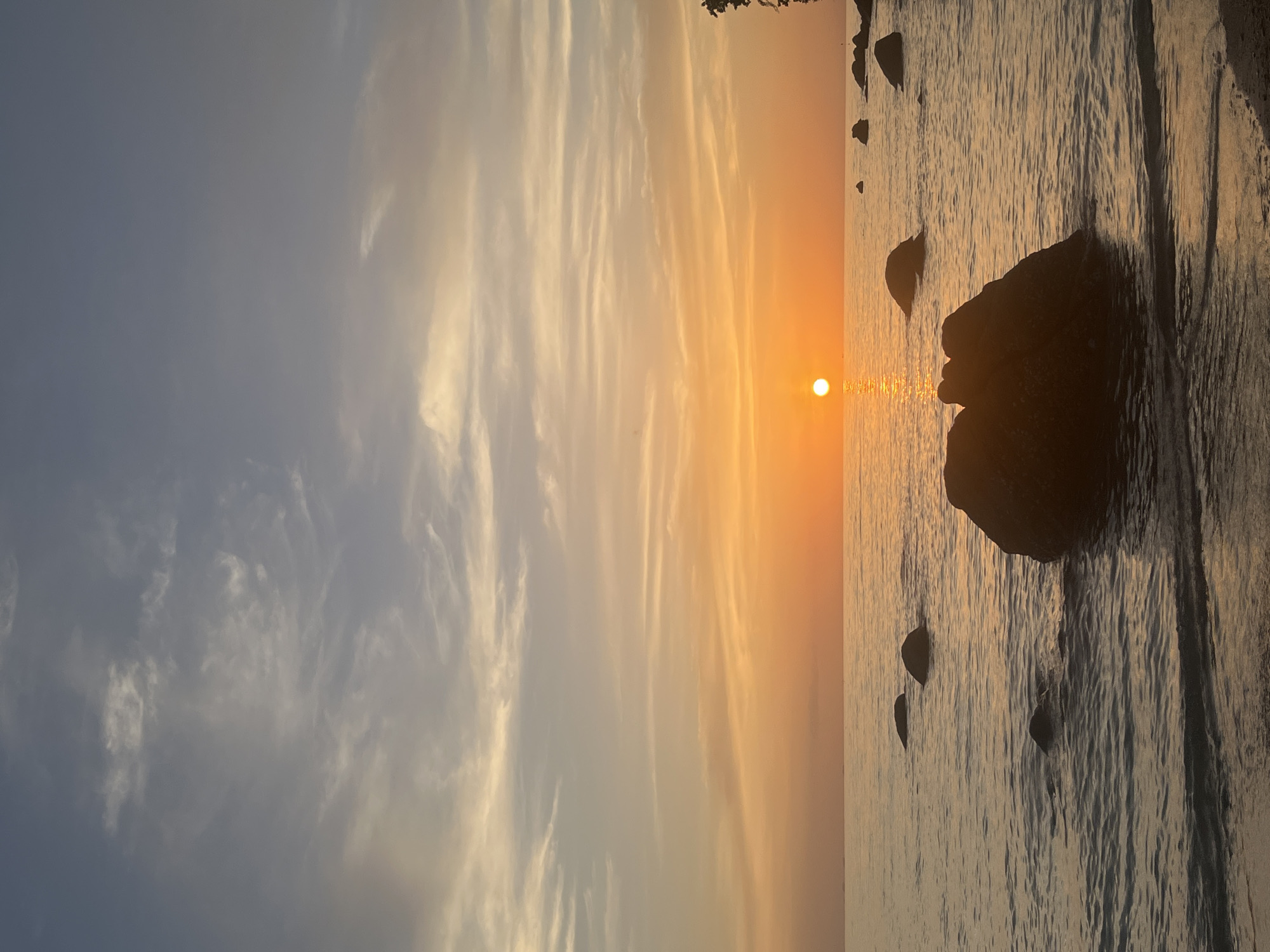 A serene sunset scene is captured on a beach in Ko Pha-ngan District, Thailand. The sun, a bright orange orb, hovers just above the horizon, casting a warm glow across the water and sky. Wispy clouds streak across the upper portion of the sky, transitioning from a pale blue to soft shades of orange and yellow. The calm ocean reflects the vibrant colors of the sunset. Scattered rocks of various sizes emerge from the gentle waves, some partially submerged, while others rest on the sandy shore in the foreground. The overall atmosphere is peaceful and tranquil, depicting the natural beauty of a tropical beach at dusk.