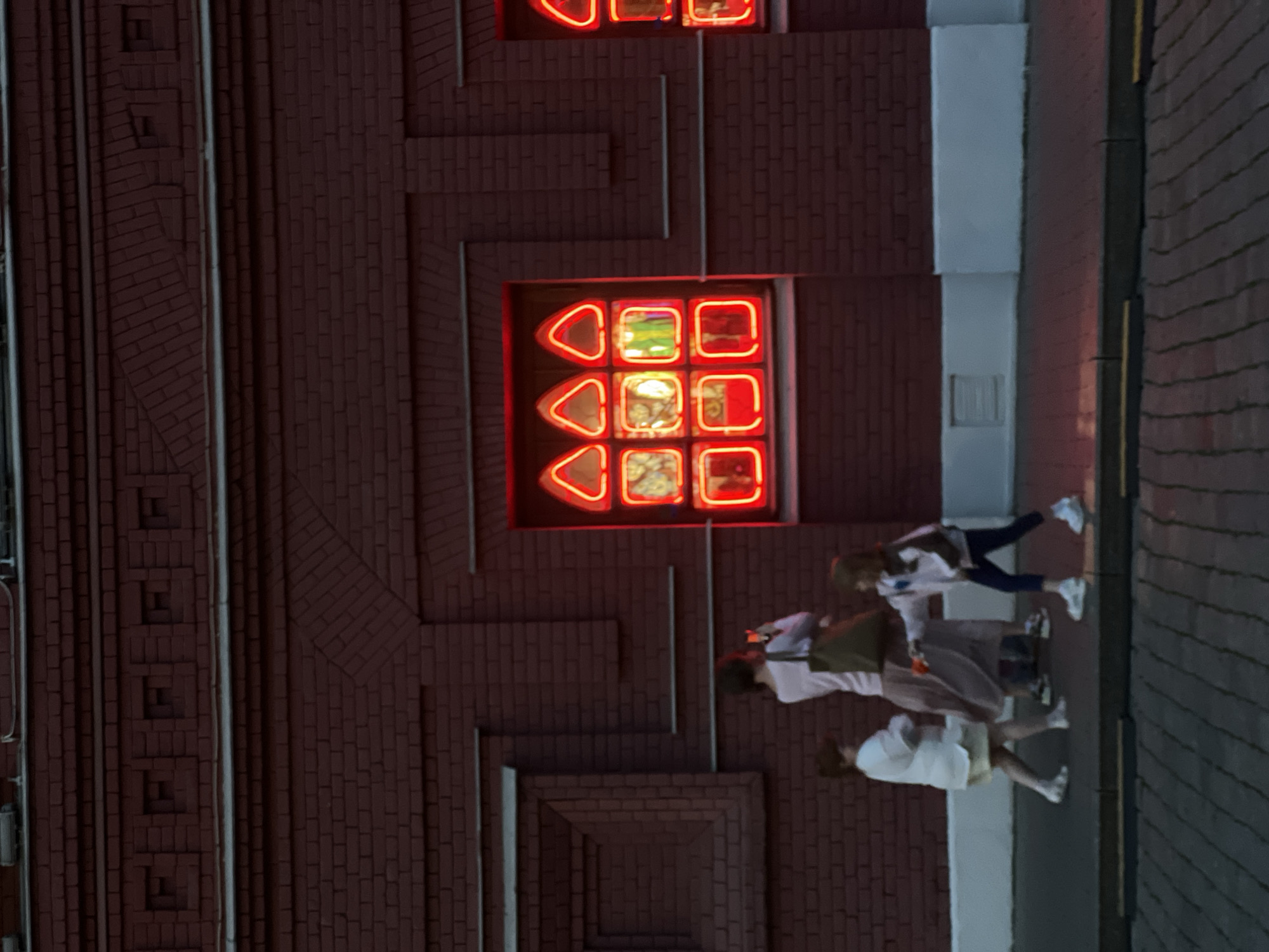 Captured at night, the image shows a brick building in Khimki, Russia. The building features a red neon display, comprised of geometric shapes, embedded in its facade. A group of people are walking on a sidewalk next to the building, appearing blurry due to the nighttime setting and probable motion. The sidewalk borders the building on one side and a street on the other. The lighting suggests an urban environment, likely illuminated by streetlights and the neon signage.