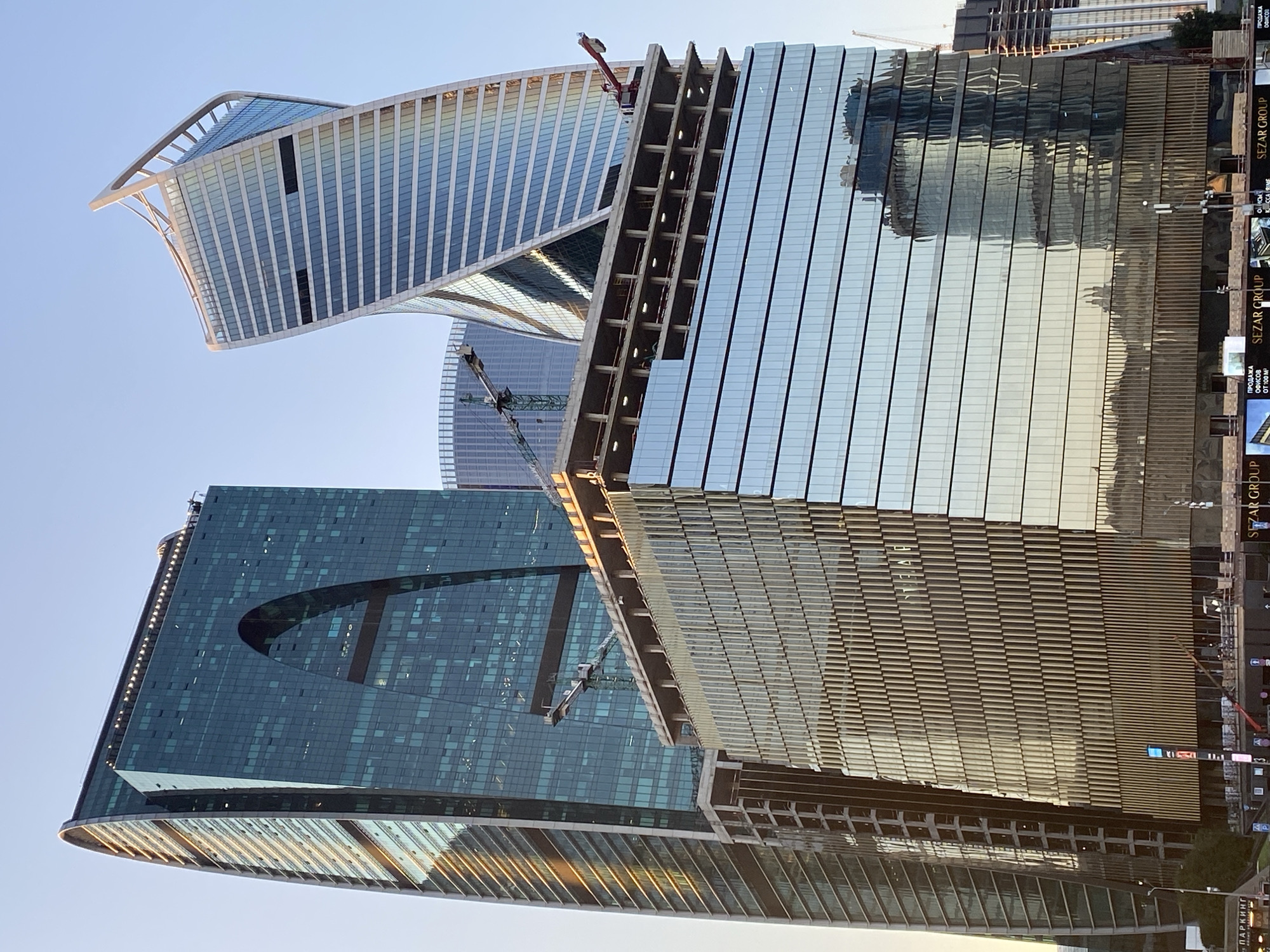 A low-angle shot shows several modern skyscrapers in an urban setting under a clear sky. The buildings are predominantly glass and steel, with one skyscraper featuring a distinctive archway cut into its facade. Another skyscraper has a twisted, aerodynamic shape, tapering as it rises. In the foreground, a partially constructed building with a glass facade reflects the surrounding structures. Construction cranes are visible, indicating ongoing development. The overall impression is of a contemporary cityscape with ambitious architectural designs.