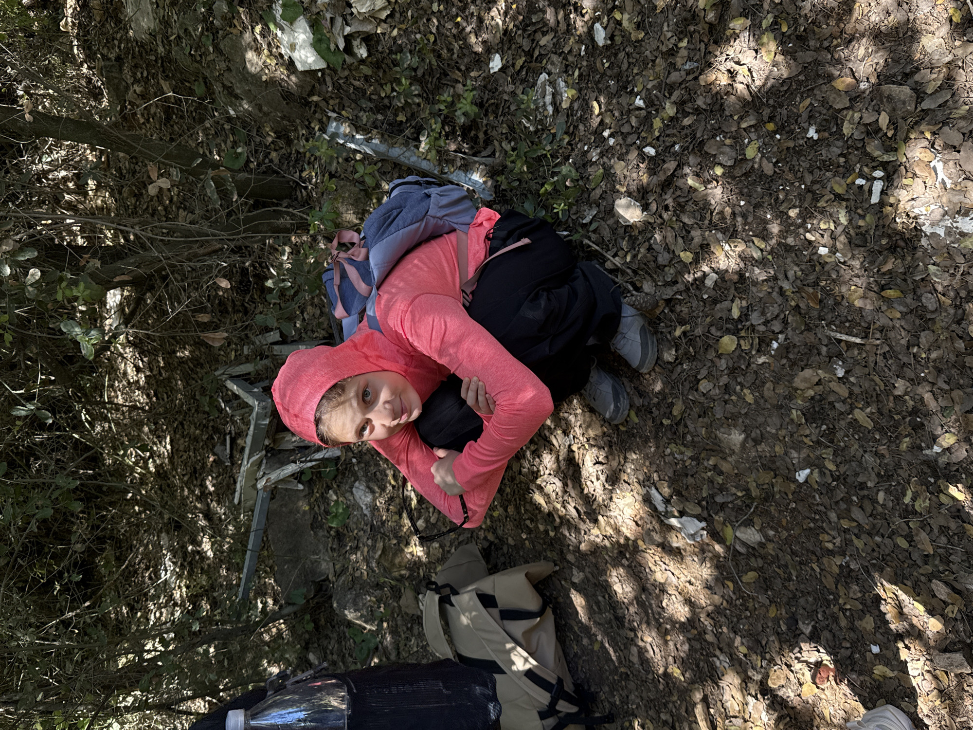An overhead photograph captures a young female individual pausing in a dry, wooded environment within the Mate Yehuda Regional Council, Israel. The person is sitting or crouching on the ground, which is covered with an abundance of brown, dry leaves, dirt, small rocks, and patches of green vegetation. She is dressed in a bright coral-pink long-sleeved hooded top, black pants, and grey athletic or hiking shoes. A blue and pink backpack is worn on her back, and a tan-colored bag or backpack rests on the ground near her. A clear water bottle is also visible nearby.
She looks directly up towards the camera with a calm, neutral expression. The setting features dappled sunlight filtering through trees, casting distinct shadows on the ground, indicating a bright, sunny daytime. In the background and mixed with the leaf litter, there are scattered pieces of white debris, possibly plastic or other litter, and some metallic-looking fragments. The scene suggests a break during an outdoor activity like hiking or exploring. There is no visible text.