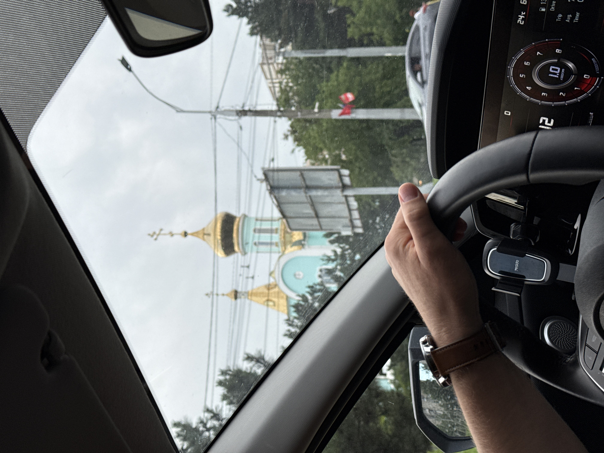 The image shows a person's hands on the steering wheel of a car, driving through a city street. The car's dashboard is visible on the right, displaying speed and other information. In the background, a blue and gold domed church is visible, along with trees and utility poles. The sky is overcast.