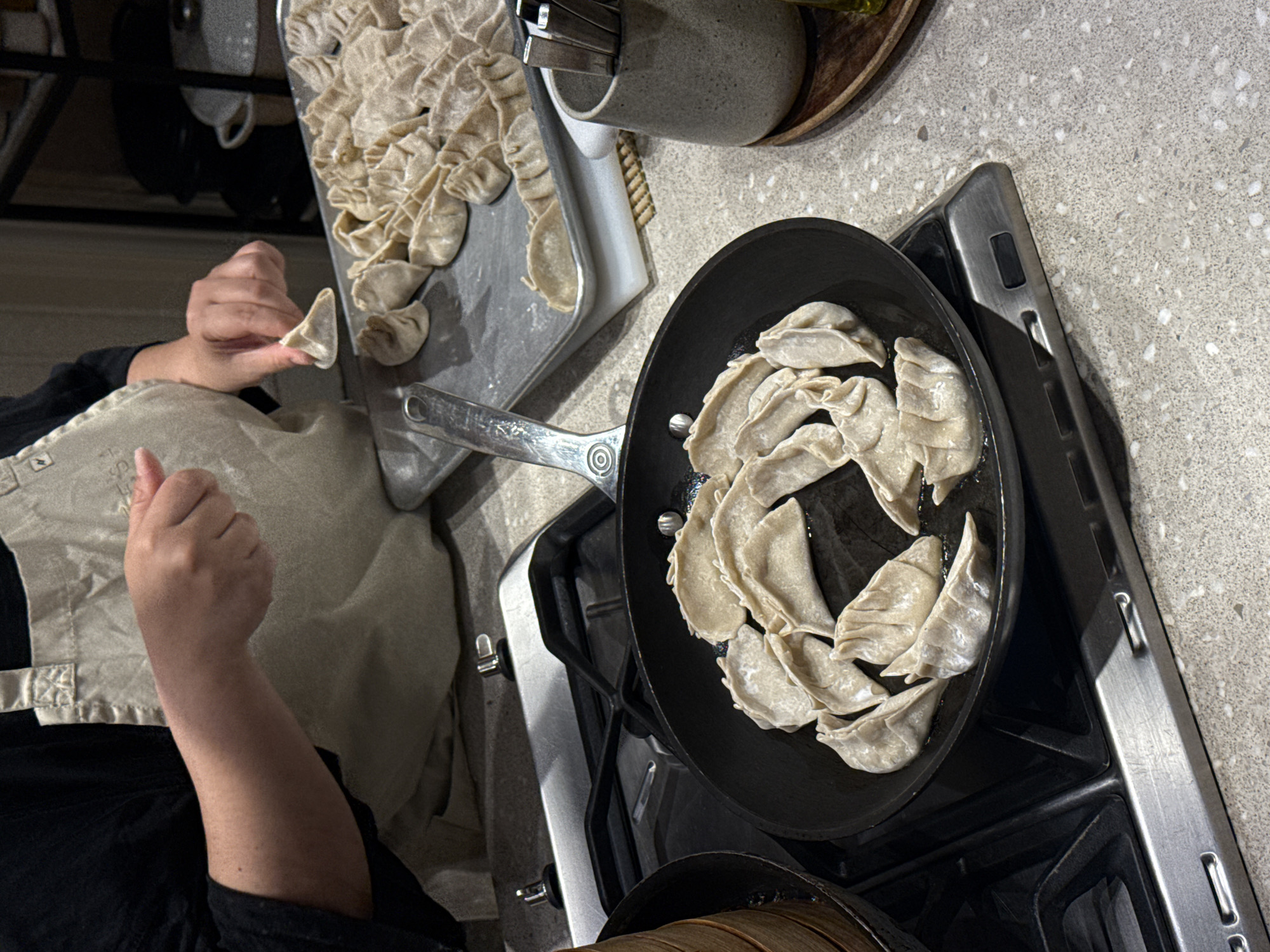 A person wearing an apron is preparing dumplings in a kitchen. A batch of uncooked dumplings is on a baking sheet next to a frying pan on a stovetop, where another batch of dumplings is frying in oil. Utensils are in a container next to the baking sheet. The location is likely a residential kitchen, and there is no verifiable evidence to indicate the city of Mexico City, Mexico.