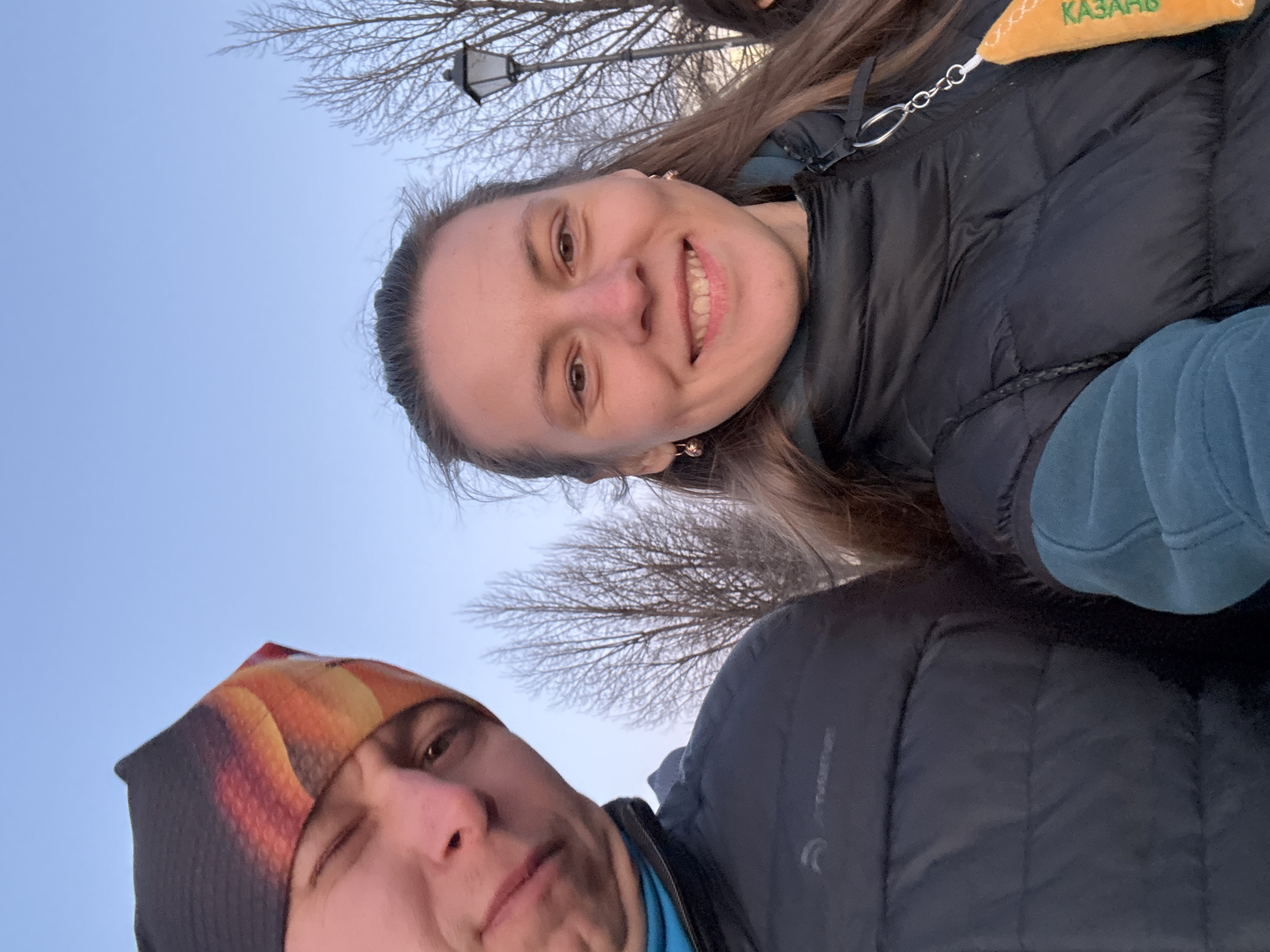 The image shows two people in an outdoor setting, seemingly taking a selfie against a light blue sky with a few bare tree branches visible. One person is wearing a colorful beanie and a blue shirt, winking at the camera. The other person is smiling and wearing a dark quilted jacket with a 'Kazan' keychain attached to it. A lamppost can be seen behind them, between their heads, contributing to the contextual detail that the location might be in Kazan, Russia.