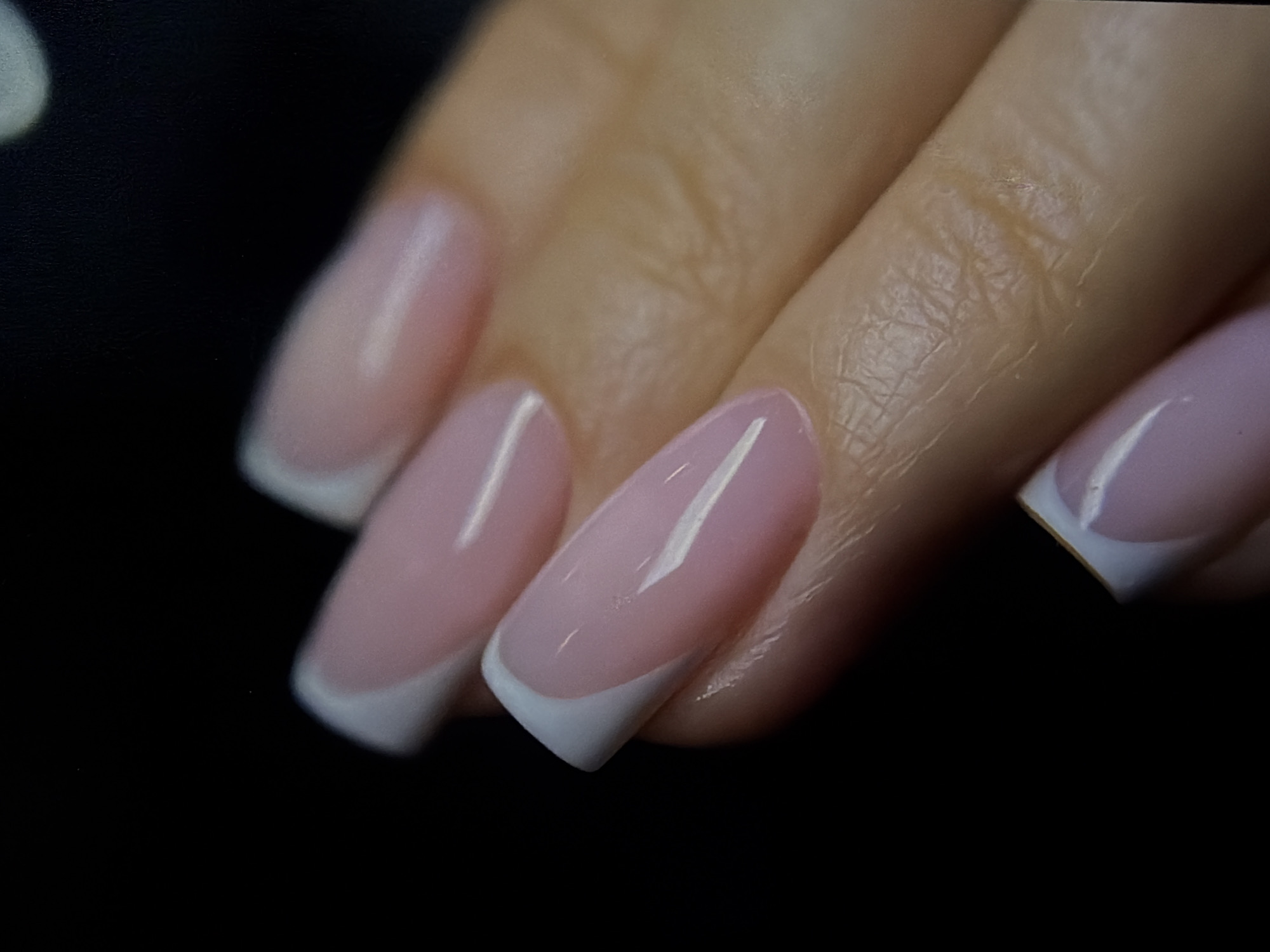 The image shows a close-up of a hand with manicured nails. The nails have a light pink base with a white French tip design. The skin is visible and shows fine lines and texture. The background is a dark, blurred area, providing a stark contrast to the hand and nails.