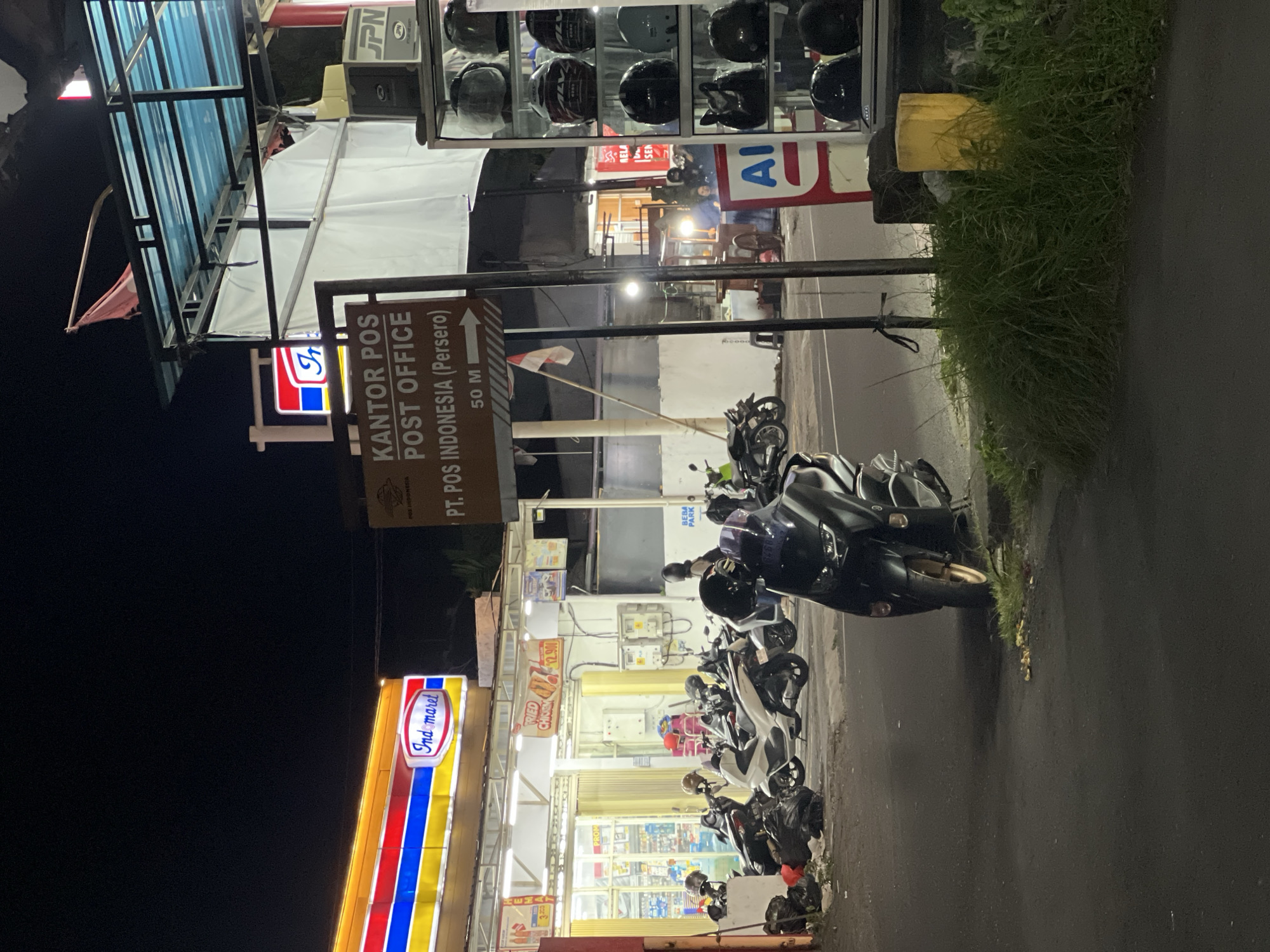 The image shows a nighttime street scene in Gianyar, Indonesia, with a focus on a convenience store and a post office. To the left, the brightly lit facade of an Indomaret convenience store is visible, featuring its characteristic red, blue, and yellow signage. In front of the store, several motorcycles are parked, some with helmets on their seats. A sign for "KANTOR POS POST OFFICE PT. POS INDONESIA (Persero)" is prominent in the center of the frame, indicating the post office's proximity. The sign is a brown rectangular board with white text and an arrow pointing to the right, suggesting a distance of 50 meters. A small Indonesian flag is also visible on a pole near the sign. To the right, a display rack filled with helmets is set up outside what appears to be a shop. The street is paved, and there is a patch of overgrown grass in the foreground. The overall atmosphere is that of a typical urban commercial area at night.