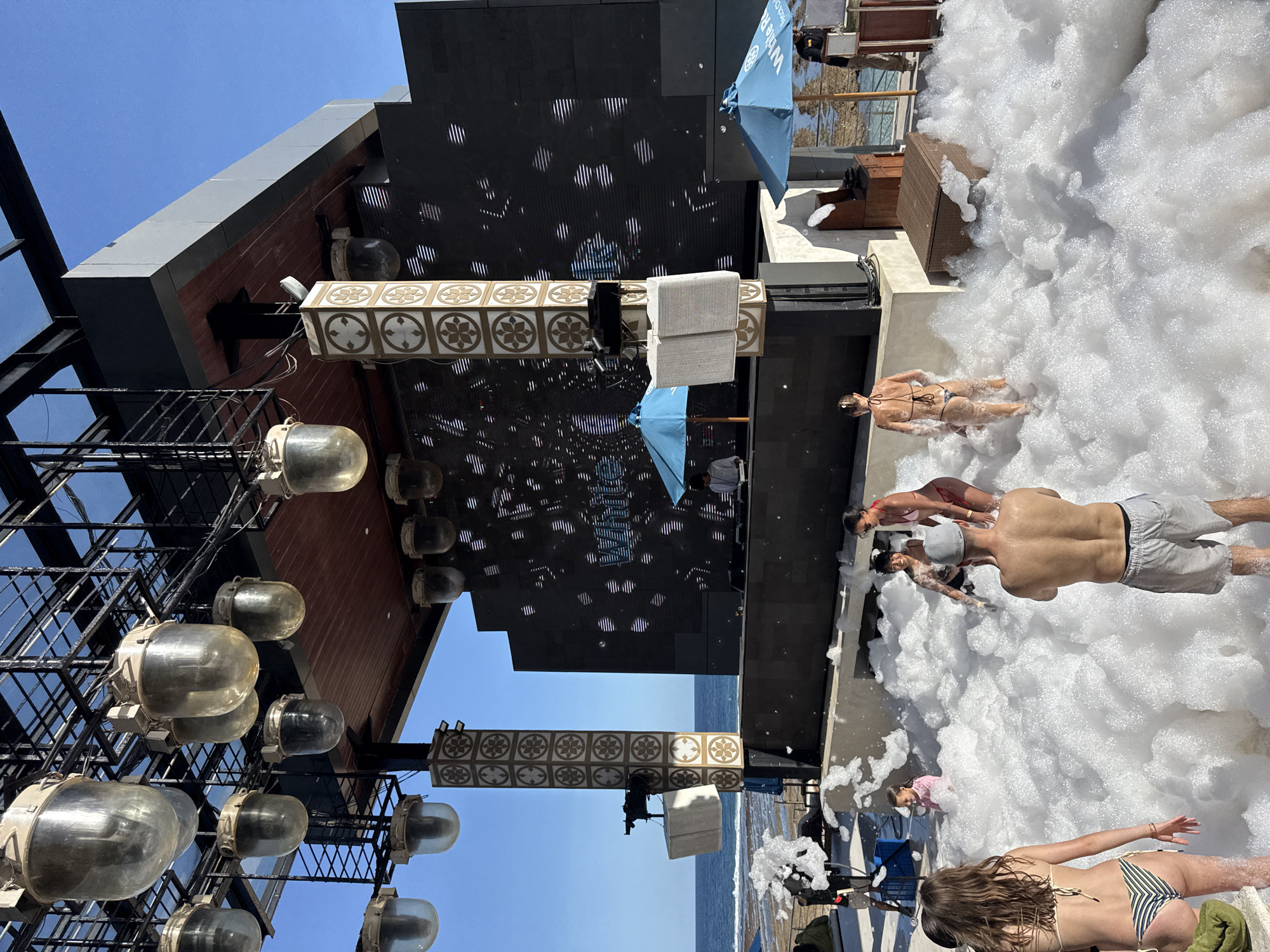 This bright, sunny image depicts an energetic outdoor foam party at a modern beach club or resort, located in Kuta Selatan, Indonesia. The scene is awash with a large quantity of white, fluffy foam covering the ground area where people are gathered.
Several individuals are visible, enjoying the foam. In the foreground and midground, people in swimwear, including women in bikinis and men in swim shorts, are wading, standing, or partially submerged in the thick bubbles. One woman with long, wavy brown hair, wearing a striped bikini, stands with her back to the camera, partially covered by the foam. Nearby, a man with a bare back and light shorts is also in the foam, along with a woman in a red bikini and other indistinct figures. Their postures and expressions suggest they are relaxed and having fun.
To the left, a contemporary building features dark paneled walls and a series of large, glass-domed industrial lights mounted vertically. Below these lights, a decorative rectangular panel with repeated floral or geometric patterns in gold and white adds a touch of elegance. A prominent large black screen, possibly an LED display, shows dynamic light patterns. Two light blue umbrellas are visible; one in the upper middle clearly shows the partial text "White Be".
The background reveals a clear, bright blue sky and the expanse of a calm blue ocean, reinforcing the coastal setting. Hints of dark outdoor furniture, like lounge chairs, can be seen amidst the foam or on the surrounding deck. The overall impression is one of a lively daytime event, combining modern aesthetics with a fun, resort-style atmosphere.