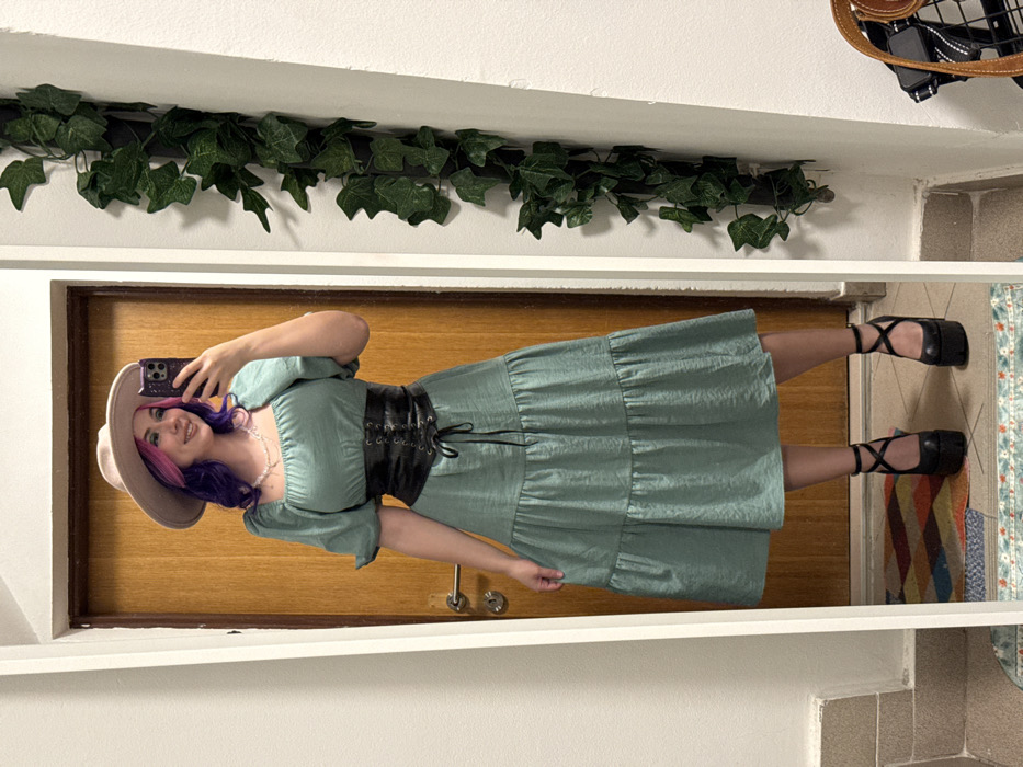 A woman in a green dress with puffy sleeves and a corset belt stands in front of a mirror, taking a selfie. She's wearing a beige hat and black platform shoes with cross straps. Her hair is purple with a pink streak. Artificial ivy decorates the top of the mirror frame. The reflection shows a wooden door and a colorful rug on the floor. A bag is visible in the top corner. The overall setting appears to be inside a house.