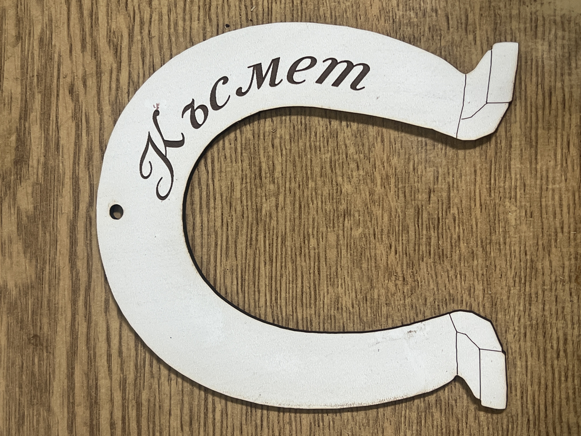 A white horseshoe with Cyrillic letters on it is placed on a wood-grained surface. The letters are brown and appear to spell out a word. The horseshoe has a small hole on the left side and geometrical designs on the right side.
The word on the horseshoe is неясен.