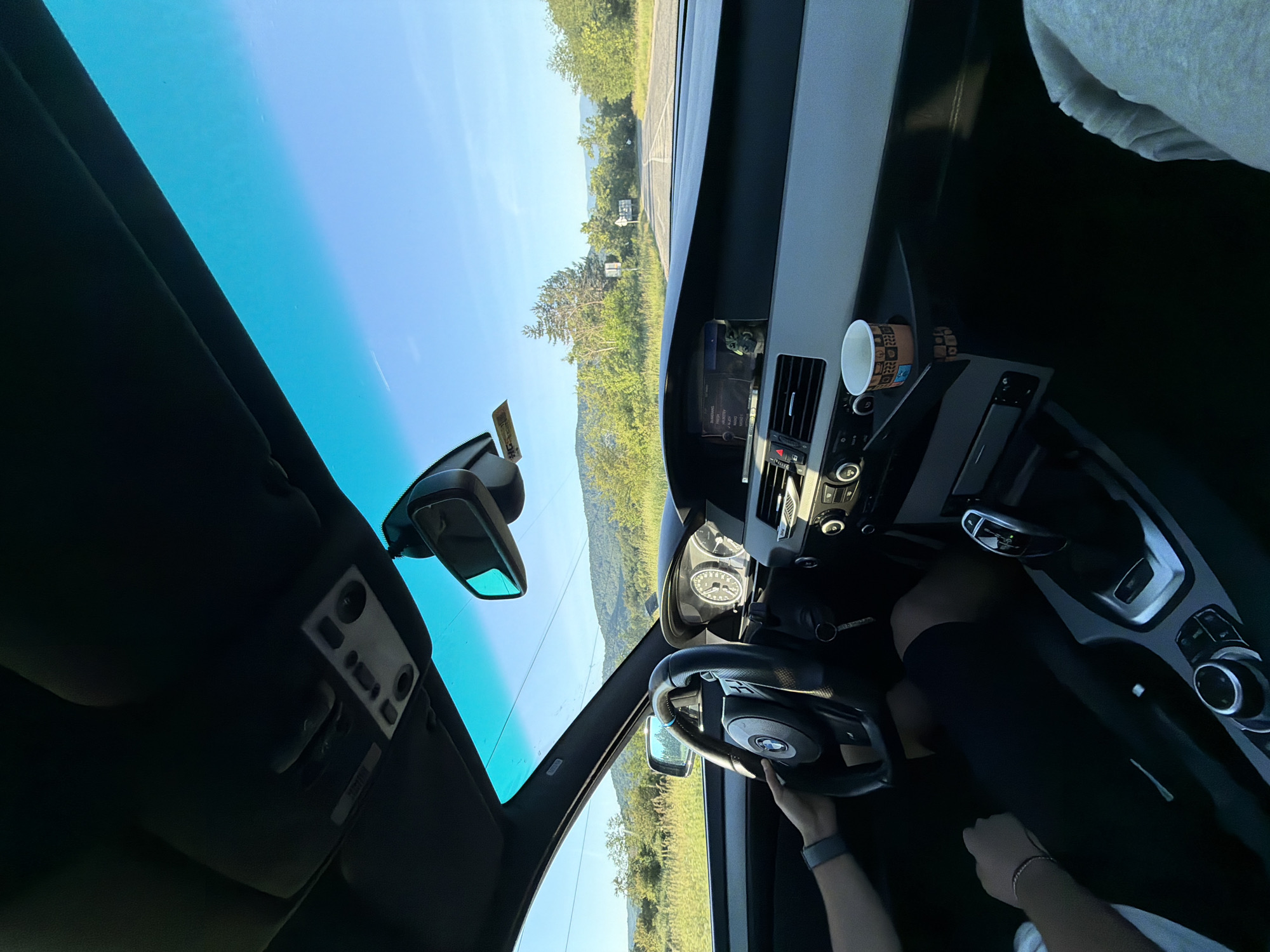 The image depicts the interior of a car, with a person's hands visible on the steering wheel. The view looks forward through the windshield, showing a road winding through a landscape of rolling hills and trees under a clear blue sky. The dashboard of the car is visible, including the steering wheel, gear shift, and a cup holder with a paper cup. The car appears to be in motion on a road outside of the city of Хаджийско, Bulgaria, given the presence of natural scenery and the absence of urban structures. The time of day appears to be late afternoon or early evening, as indicated by the sunlight casting shadows and illuminating the landscape.