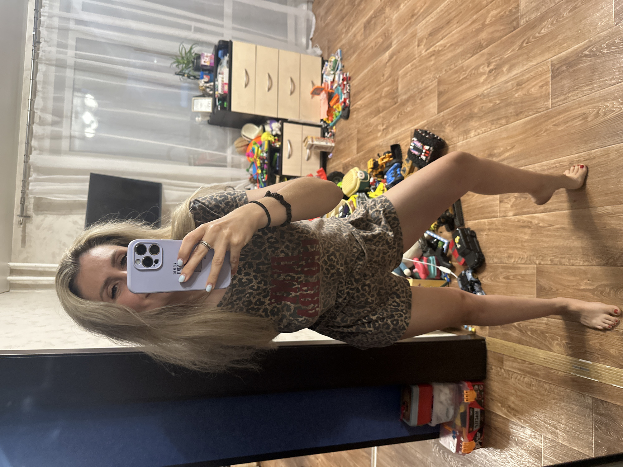 In Novoaltaysk, Russia, a young woman with blonde hair takes a mirror selfie in a room that appears to be a child's bedroom or playroom. She is wearing a leopard-print pajama set, consisting of a short-sleeved top and shorts. Her nails are painted a light blue color, and she is wearing a ring on her right hand. She is holding a purple phone in her right hand. The room is cluttered with children's toys, including various construction vehicles, building blocks, and other play items scattered across the wooden floor. A dresser with several drawers is visible in the background, as is a dark blue cabinet on the left. A television is mounted on the wall. The overall environment suggests a lived-in, perhaps chaotic, domestic setting.