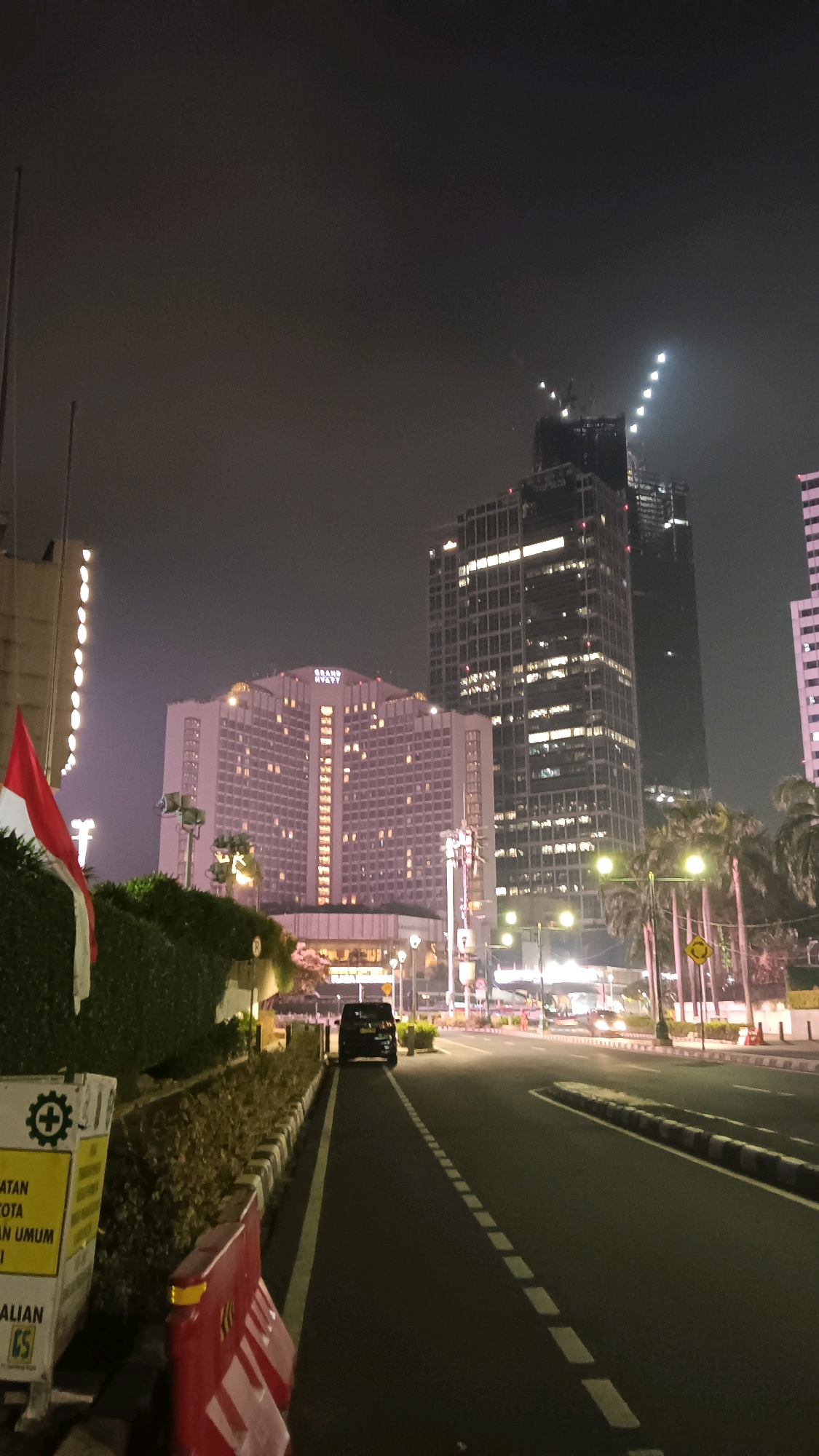 This is a nighttime, vertical, eye-level shot of a city street in Central Jakarta, Indonesia. The foreground features a dark asphalt road with dashed white lane markings. A black SUV is parked on the left side of the road, facing away from the viewer. To the left of the road, there is a row of decorative plants and a red and white construction barrier. Behind the barrier, a yellow sign with text in Indonesian is visible. Further left, a portion of a green hedge is present, and a red and white flag is partially visible.
In the midground, a large, illuminated hotel building, identified as the Grand Hyatt, dominates the scene. It is a multi-story structure with numerous lit windows. Adjacent to it, to the right, are two tall, modern skyscrapers. The building on the far right appears to be under construction, with visible scaffolding and crane structures. The sky is dark and hazy, with some lights visible in the distance. Streetlights and building lights illuminate the scene, creating reflections on the road surface. Palm trees are visible along the roadside on the right. The overall atmosphere is urban and bustling, typical of a city at night.