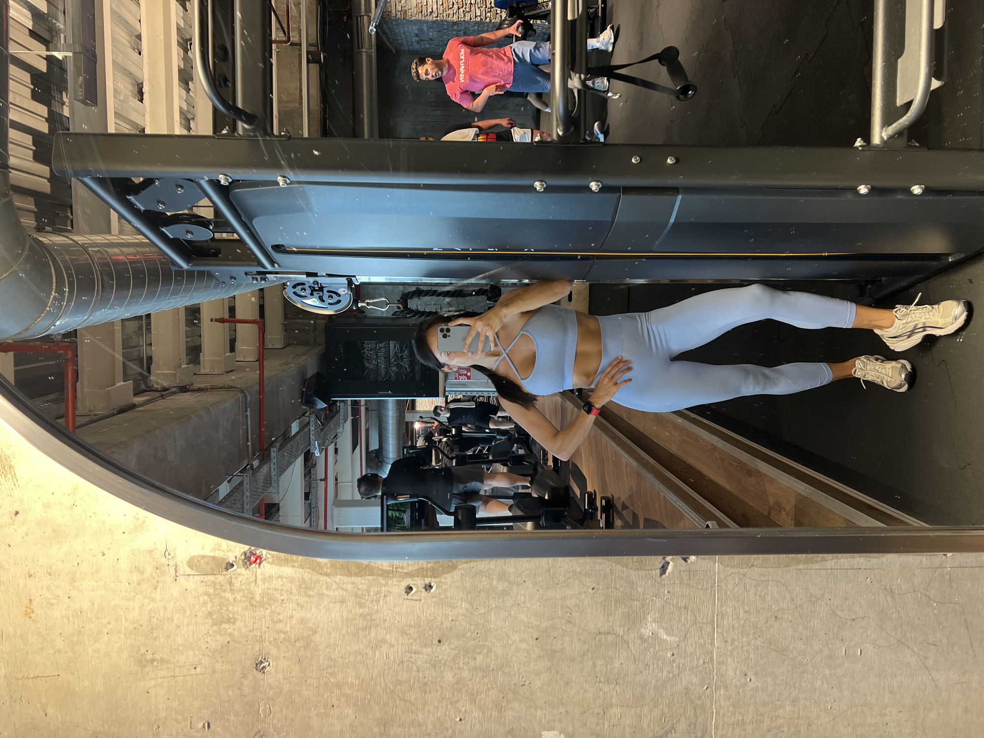 A woman is taking a mirror selfie in a gym. She is wearing a light blue sports bra and leggings. The gym has exposed ceilings with pipes and lighting fixtures, concrete walls, and wooden floors. In the reflection, other gym-goers can be seen using exercise equipment. One man in a red t-shirt and blue shorts is standing near weight racks, and another man is on a treadmill. The woman is holding her phone up to take the photo, and her left hand is on her hip. The gym appears to be a modern, industrial-style space.