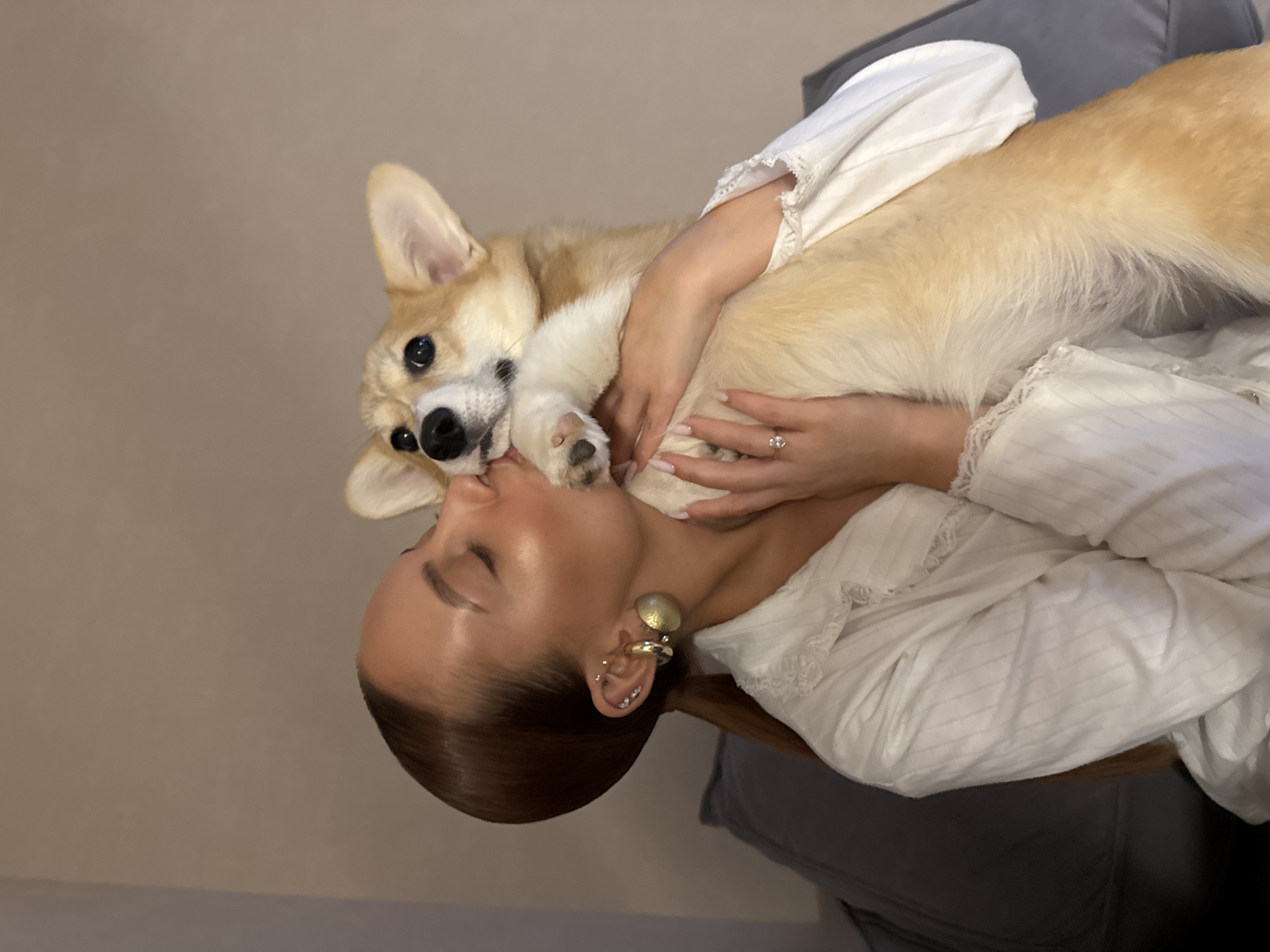 A woman in a white top holds a light brown and white corgi dog. The woman is fair-skinned with dark brown hair pulled back in a ponytail. She has multiple ear piercings and a large gold earring. Her left hand is visible and she wears a ring on her ring finger. The corgi is positioned with its paws near the woman's face. The background consists of a light beige wall and a gray couch. The location cannot be verified.
