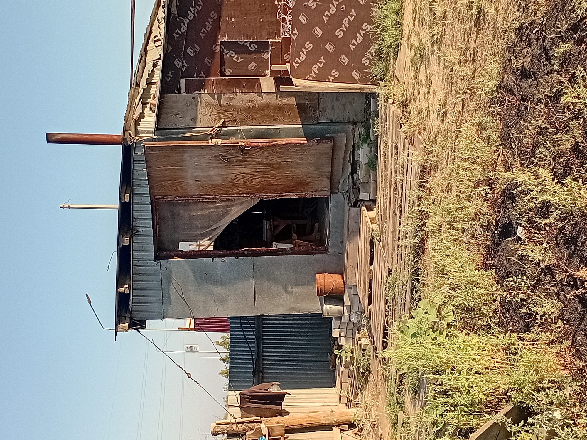 The image shows a dilapidated outbuilding, likely a shed or small workshop, constructed from corrugated metal and salvaged wood. The structure appears to be in a state of disrepair, with warped wooden doors and exposed metal sheeting. A rusty metal pipe protrudes from the roof, serving as a chimney. In front of the building, stacks of wooden pallets and some bricks are visible, along with an orange bucket and a silver bucket. The ground is covered in dry grass and dirt. In the background, a corrugated metal fence and some power lines can be seen. The overall scene suggests an outdoor, possibly rural or semi-industrial setting in the city of Semey, Kazakhstan. There are no people visible in the image.