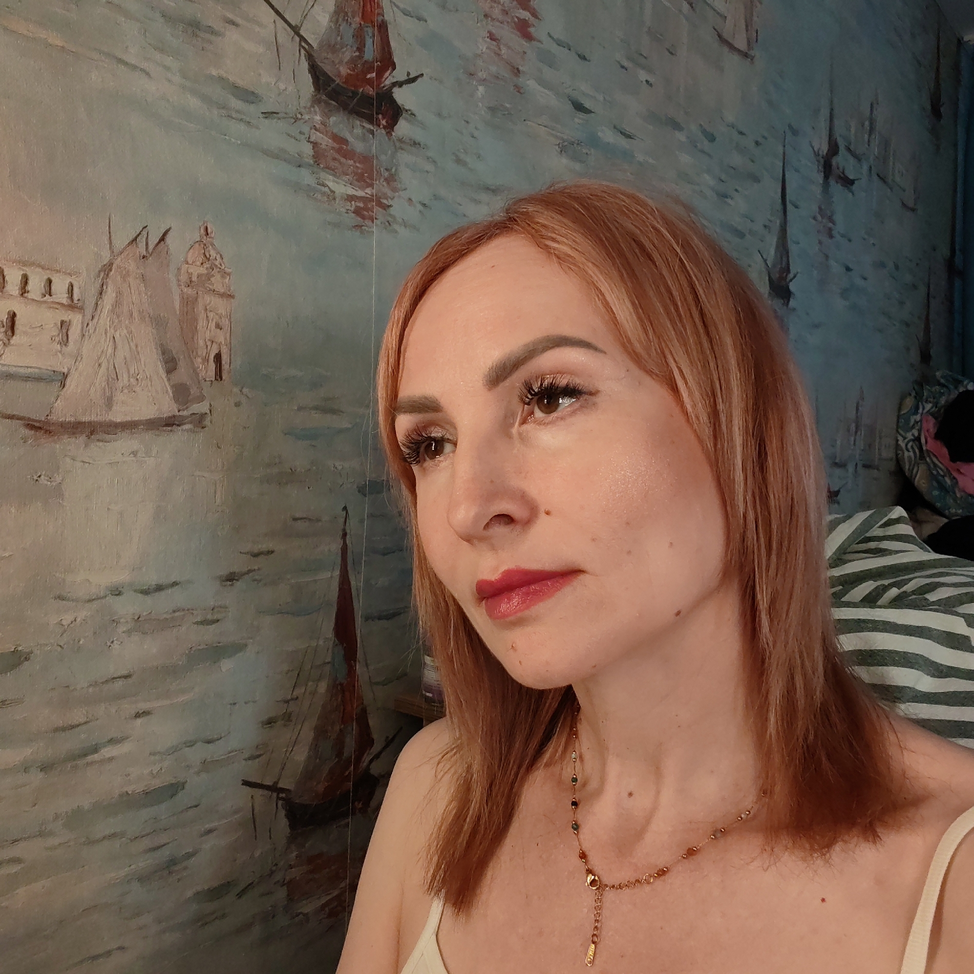 A woman with reddish-blonde hair and fair skin looks off to the left. She is wearing a red lipstick, defined eyelashes, and a delicate gold necklace. She appears to be indoors, based on the striped pillow and assorted items behind her. The wallpaper behind her depicts a painted scene of buildings and boats on water. Without further information, it is not possible to connect the location to Chaikovsky, Russia.