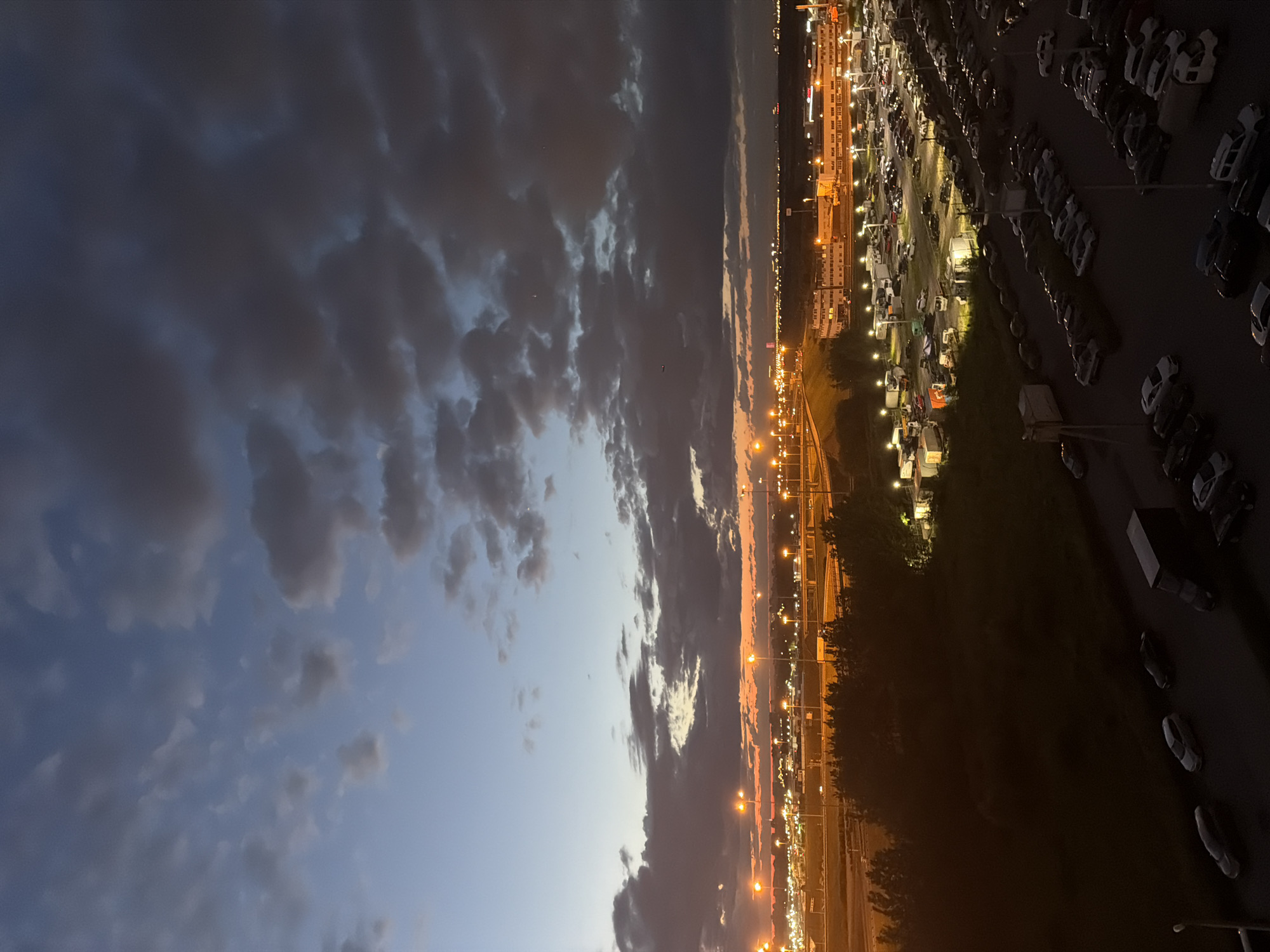 A high-angle, full shot shows a twilight scene over a vast parking lot and a cityscape. The sky is a gradient of blues and oranges, with heavy, dark clouds obscuring much of the upper portion. Below, numerous streetlights and building lights illuminate the scene. In the foreground and mid-ground, rows of parked cars are visible, mostly dark in color. Some trucks and other vehicles are also present in the parking lot. Beyond the parking lot, a complex network of roads and bridges can be seen, lit by streetlights. In the distance, a sprawling urban landscape with more lights stretches to the horizon. The overall impression is one of an active, illuminated area under a dramatic, cloudy sky at dusk.