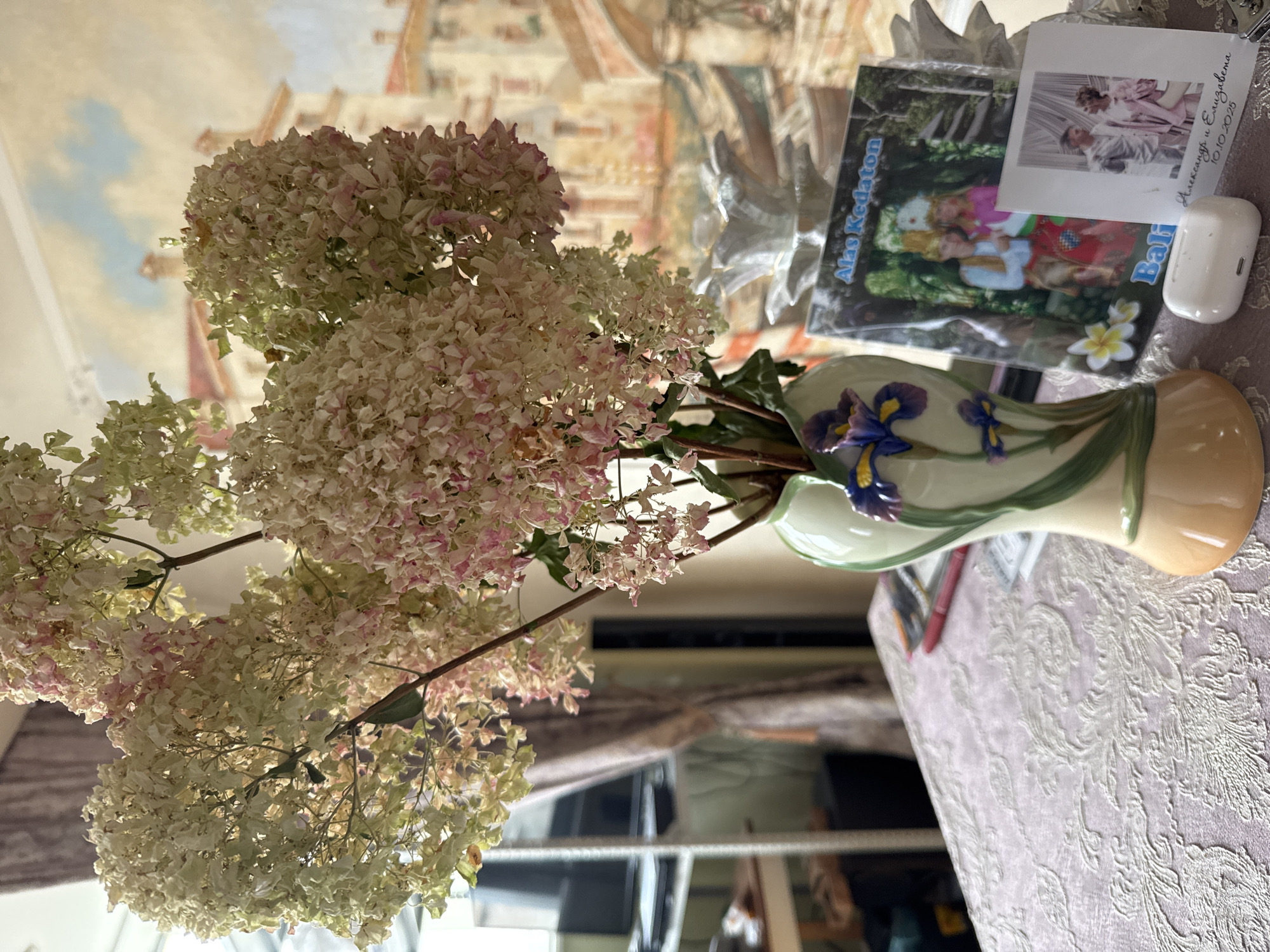 A vase of dried flowers, in shades of white and pink, sits on a patterned tablecloth on a table. To the right of the vase are two framed photographs, one larger than the other. The larger photograph shows a couple in traditional attire, with the text "Alas Kedaton Bali" above them. The smaller photograph shows a different couple, with text that appears to be names and a date below. A white wireless earbud case rests in front of the photographs. In the background, a wall mural depicts a European-style cityscape with buildings and arches. A shelf unit with electronics and a curtain are visible to the left. The scene is indoors, likely in a home. The overall lighting suggests it is daytime.