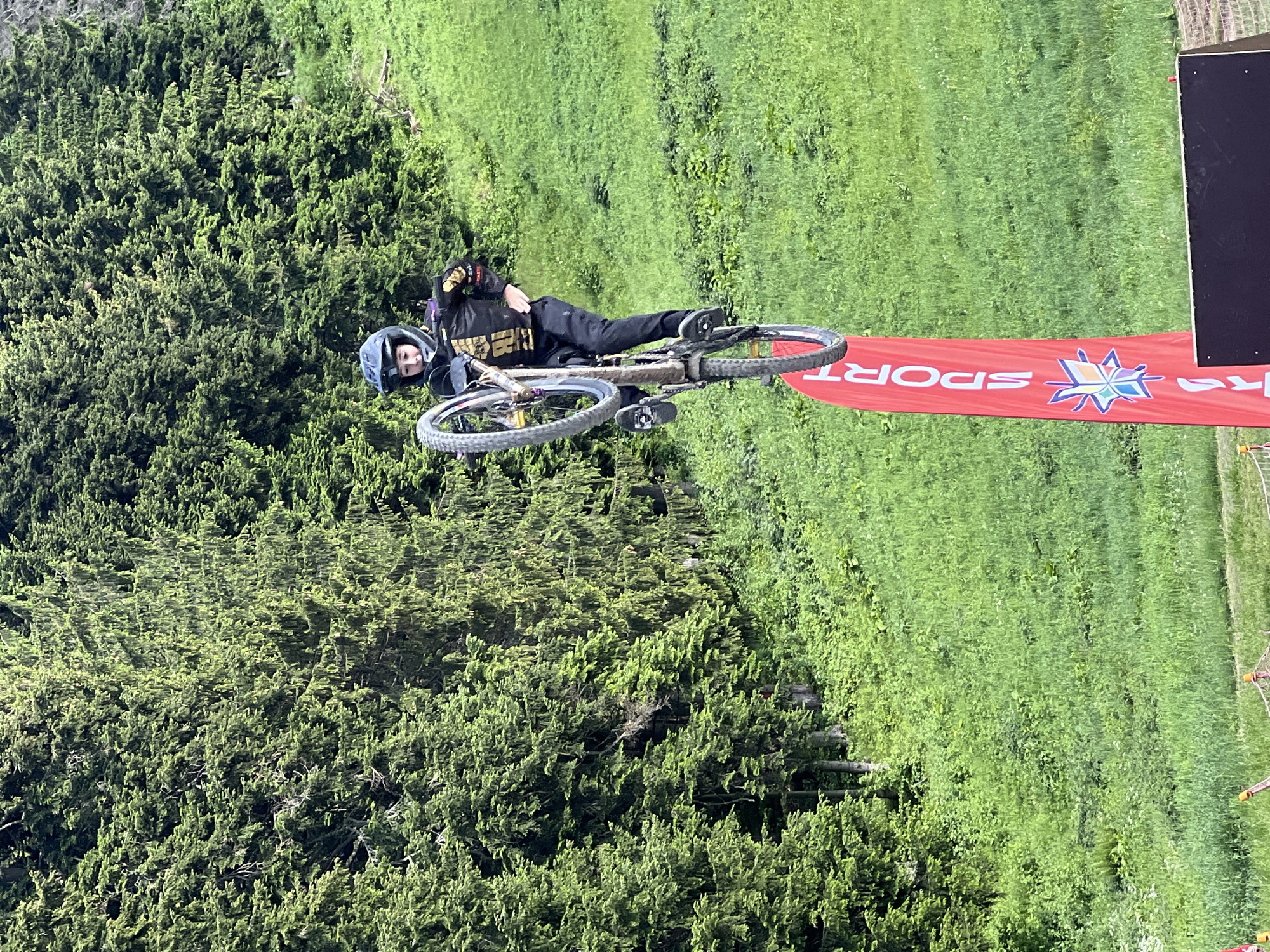 A young person wearing a helmet and protective gear is performing a bicycle stunt, with their bike in the air and tilted. They are wearing a black long-sleeved shirt with gold accents and black pants. The stunt appears to be taking place on a grassy slope with a dense forest in the background. A tall red banner with the word "SPORT" and a star logo is also visible near the bottom of the frame. The scene is outdoors in a natural environment, possibly a park or a ski resort during a warmer season.